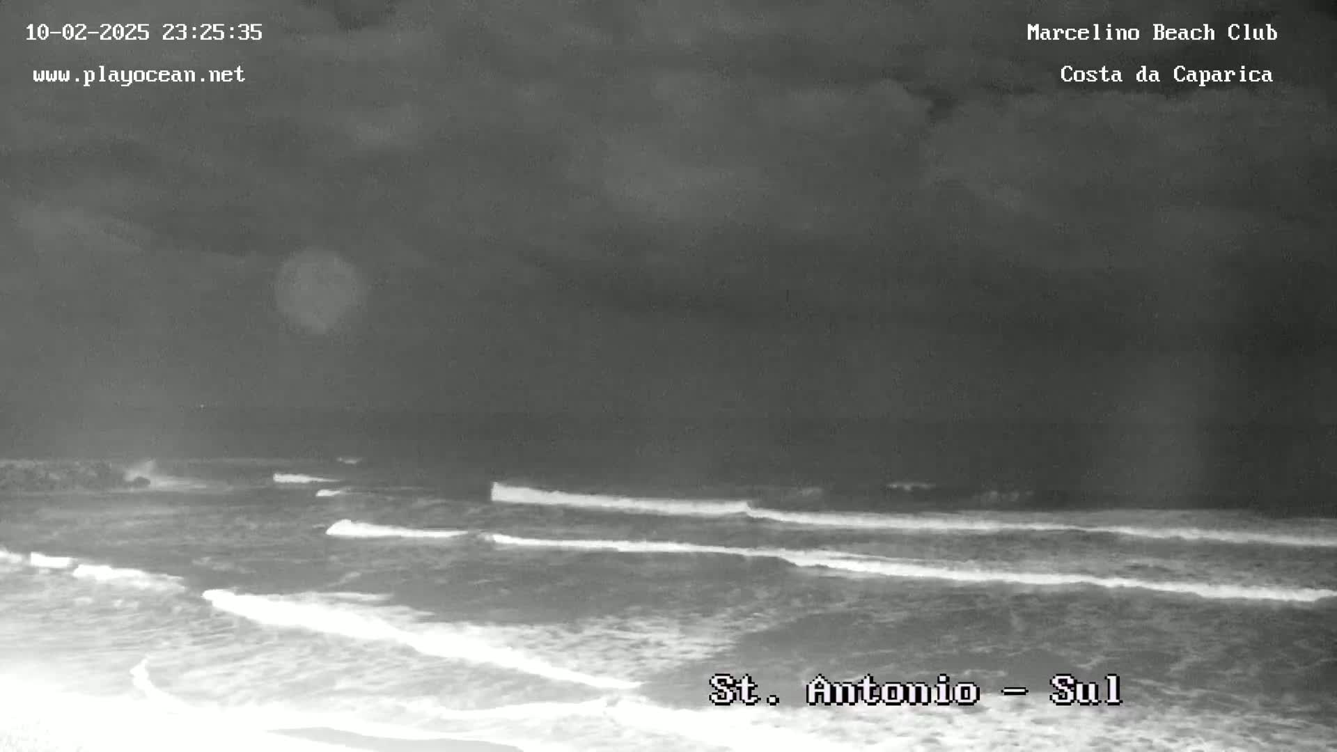 A nighttime, grayscale image shows waves breaking on a beach under a dark, cloudy sky.