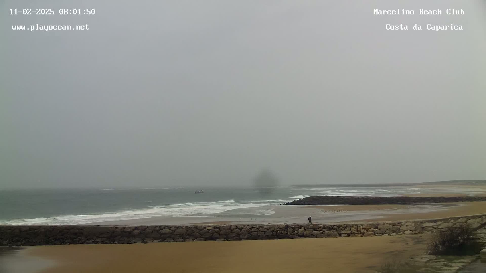 A person walks along a stone wall separating a sandy beach from a gray, overcast ocean with small waves.
