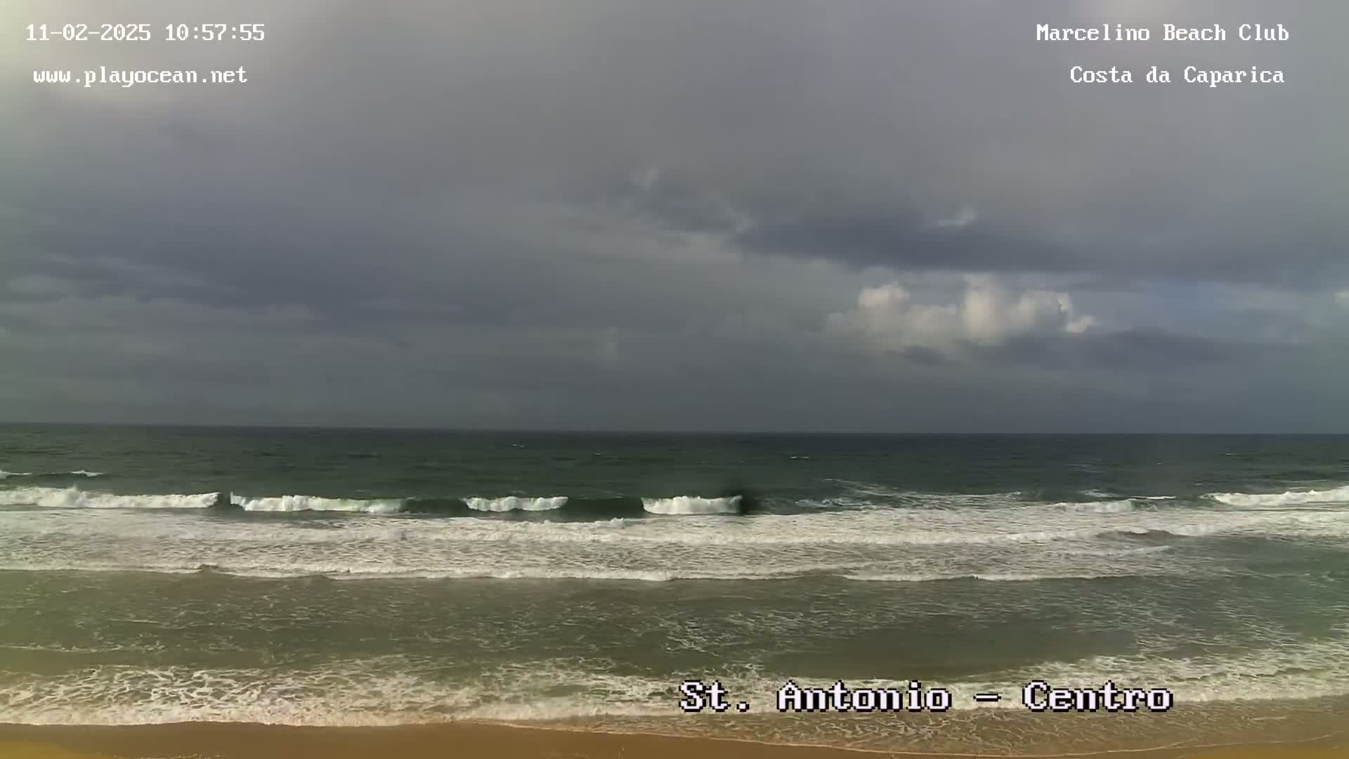 A sandy beach with moderate waves rolls into a dark, overcast ocean under a gray sky.