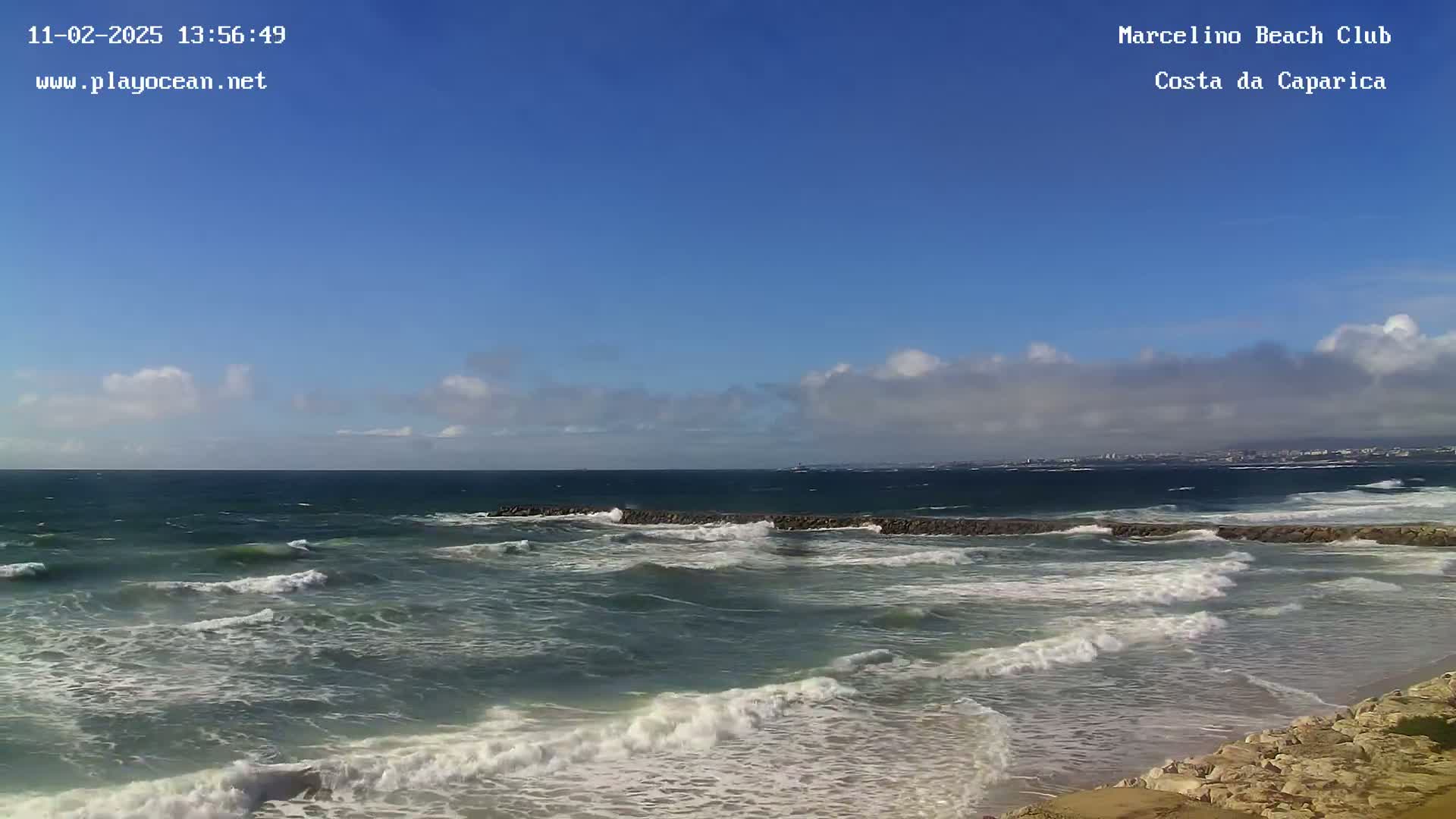 A partly cloudy blue sky overlooks a rocky shoreline with moderately sized waves crashing on the beach.