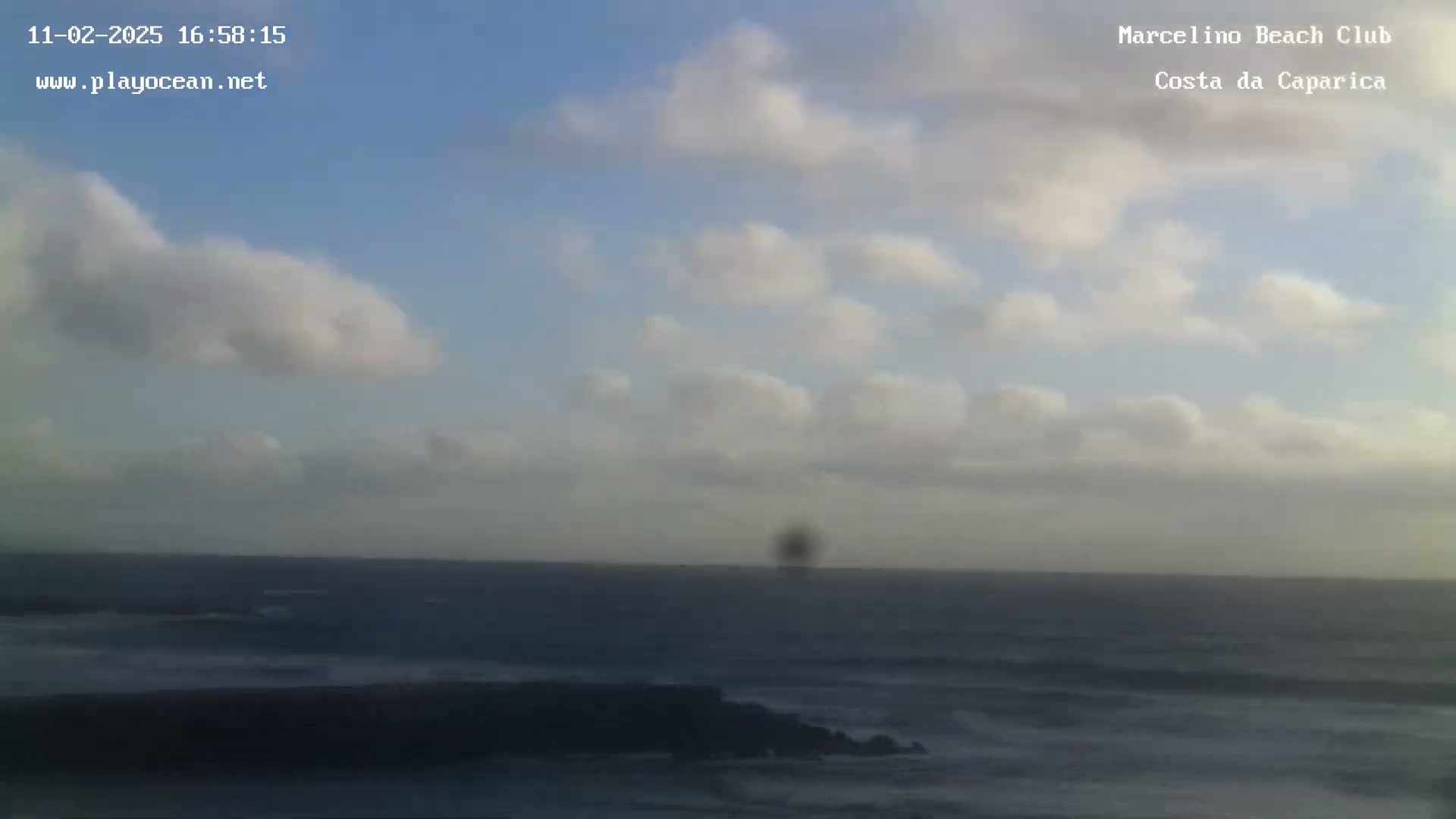 A partly cloudy sky overlooks a dark ocean with small waves breaking on a shoreline.