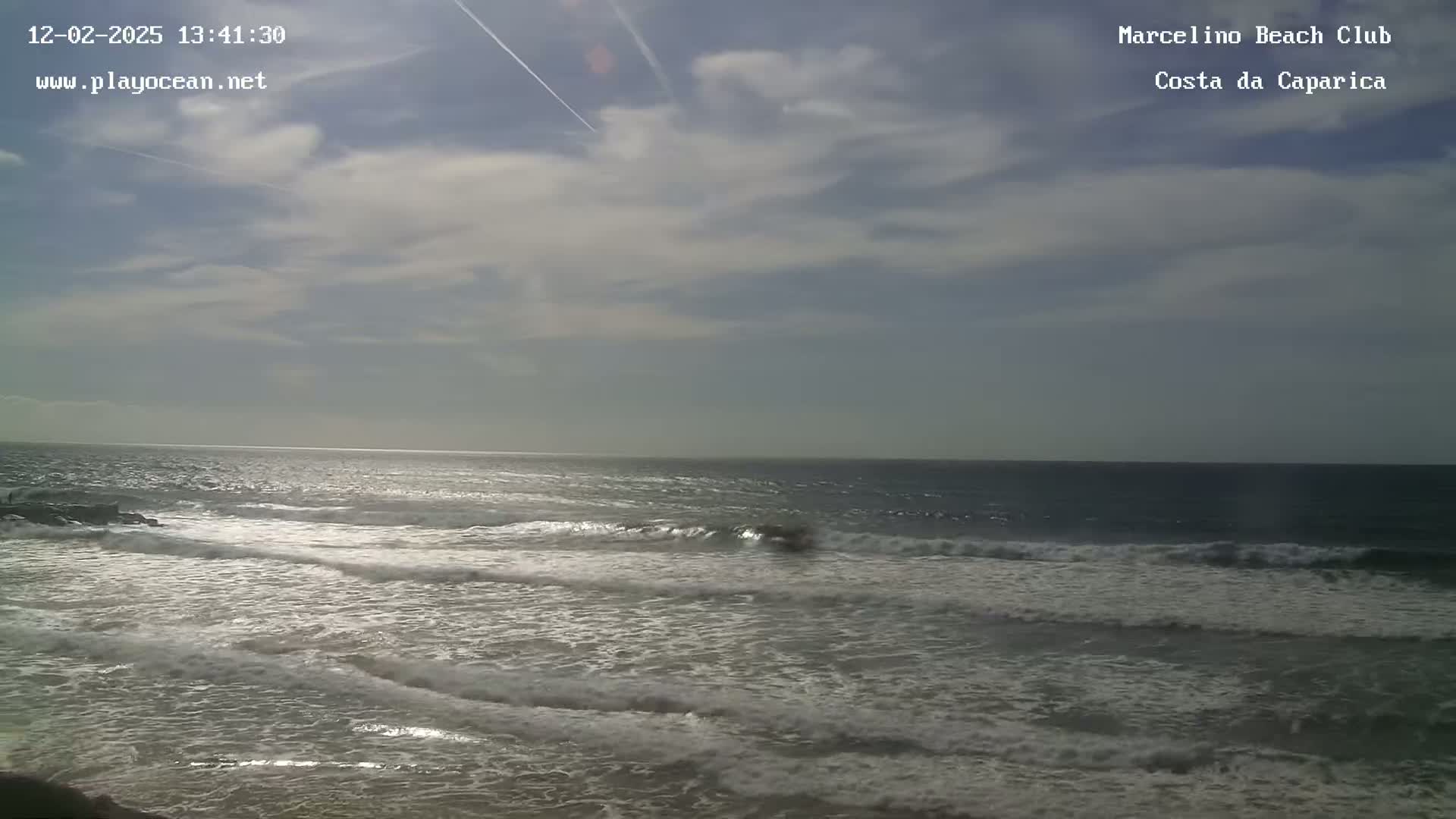 A mostly cloudy sky overlooks a gently rolling ocean with small waves breaking on the shore.
