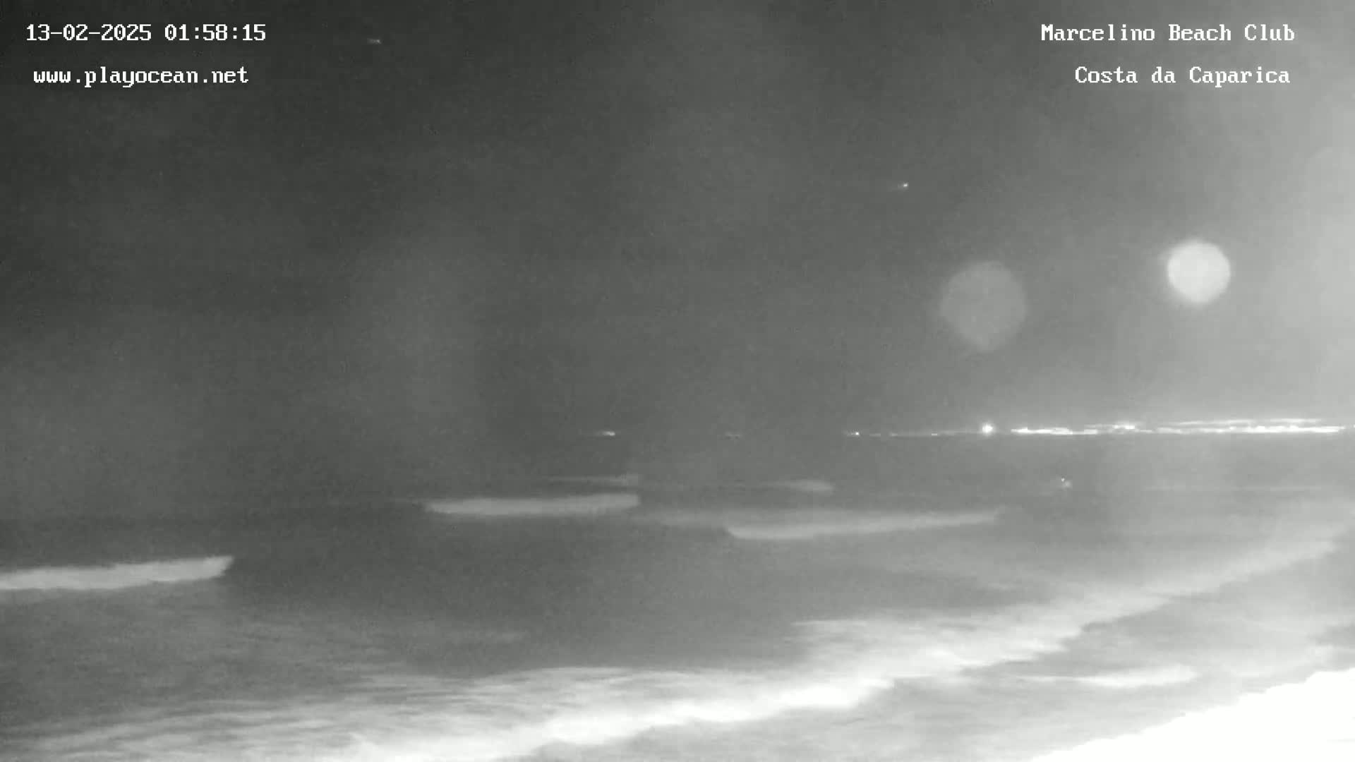 A nighttime, low-light image shows dark ocean waves lapping a shoreline under a dark sky with a few distant lights visible.