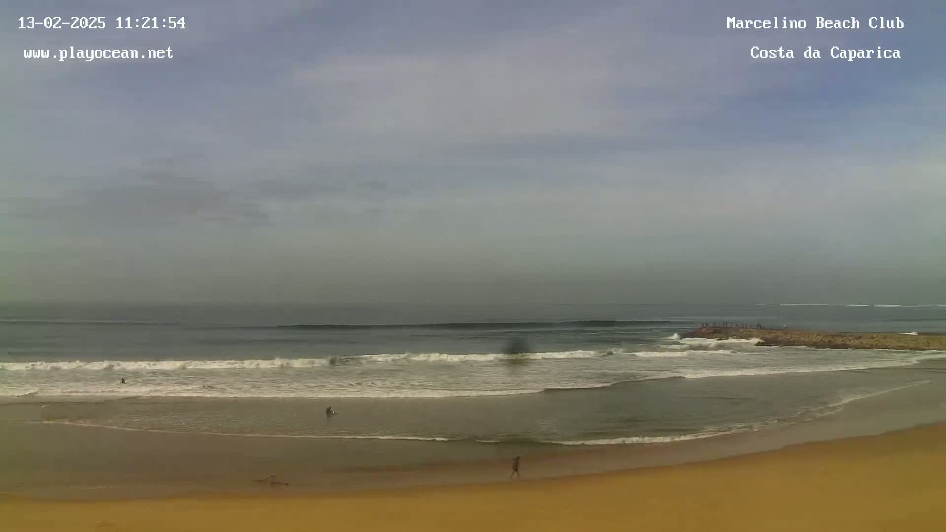 A mostly overcast sky hangs over a sandy beach with gentle waves rolling in toward a rock jetty where people are standing.