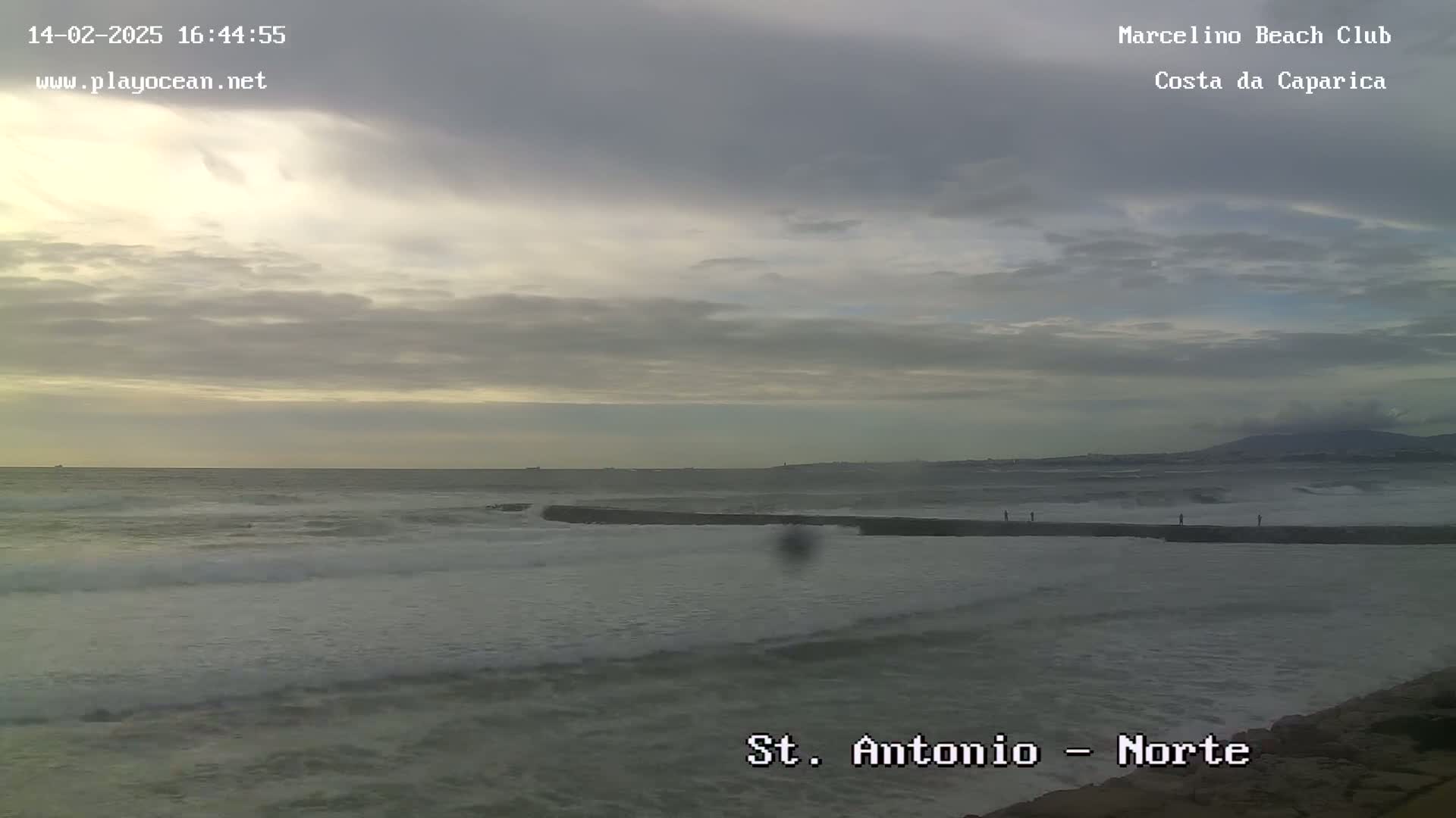 A mostly cloudy sky overlooks a rocky shoreline with moderately sized waves crashing on the beach.