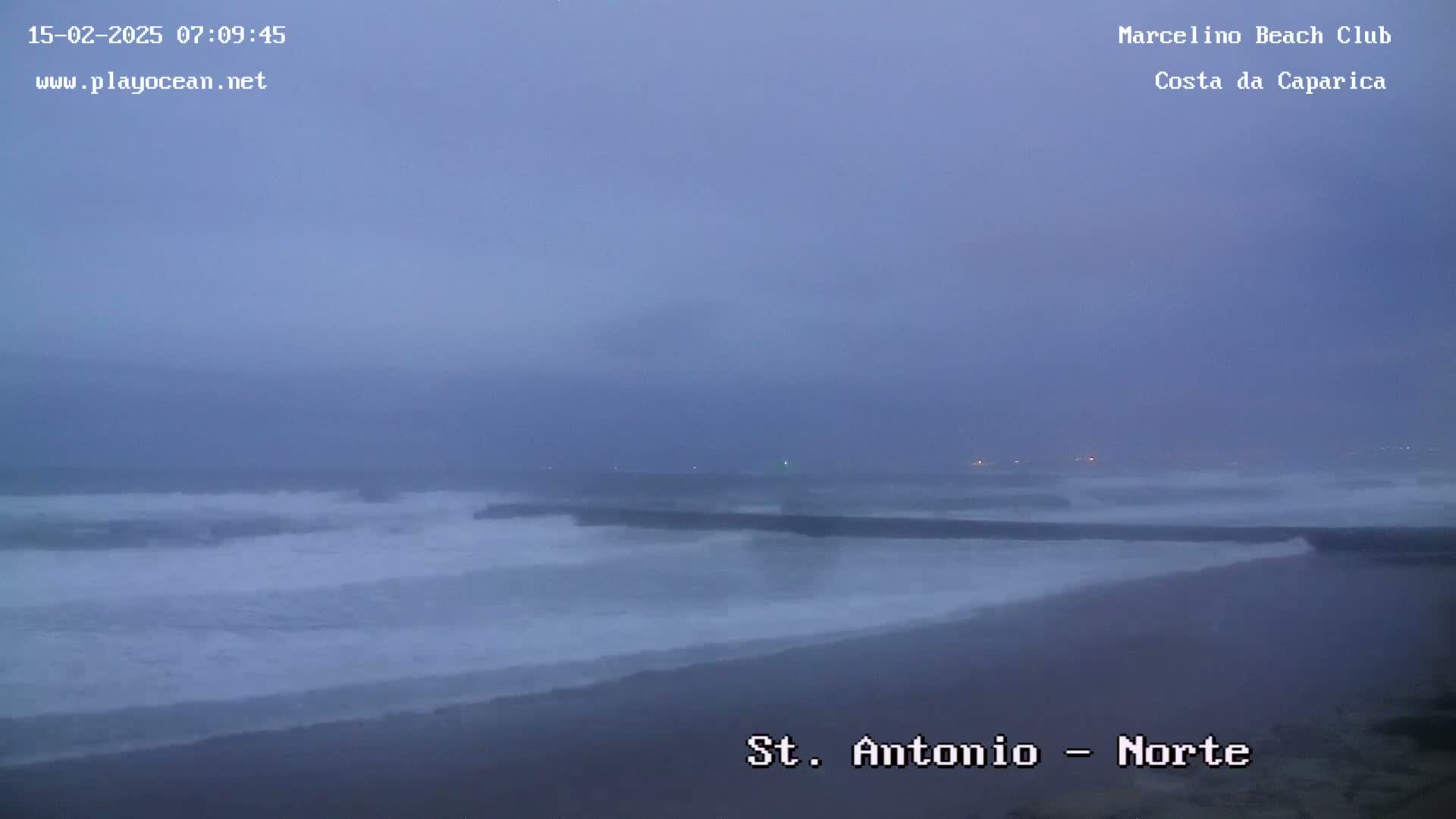 A hazy, overcast beach scene shows waves crashing against a shore with a dark structure in the water.