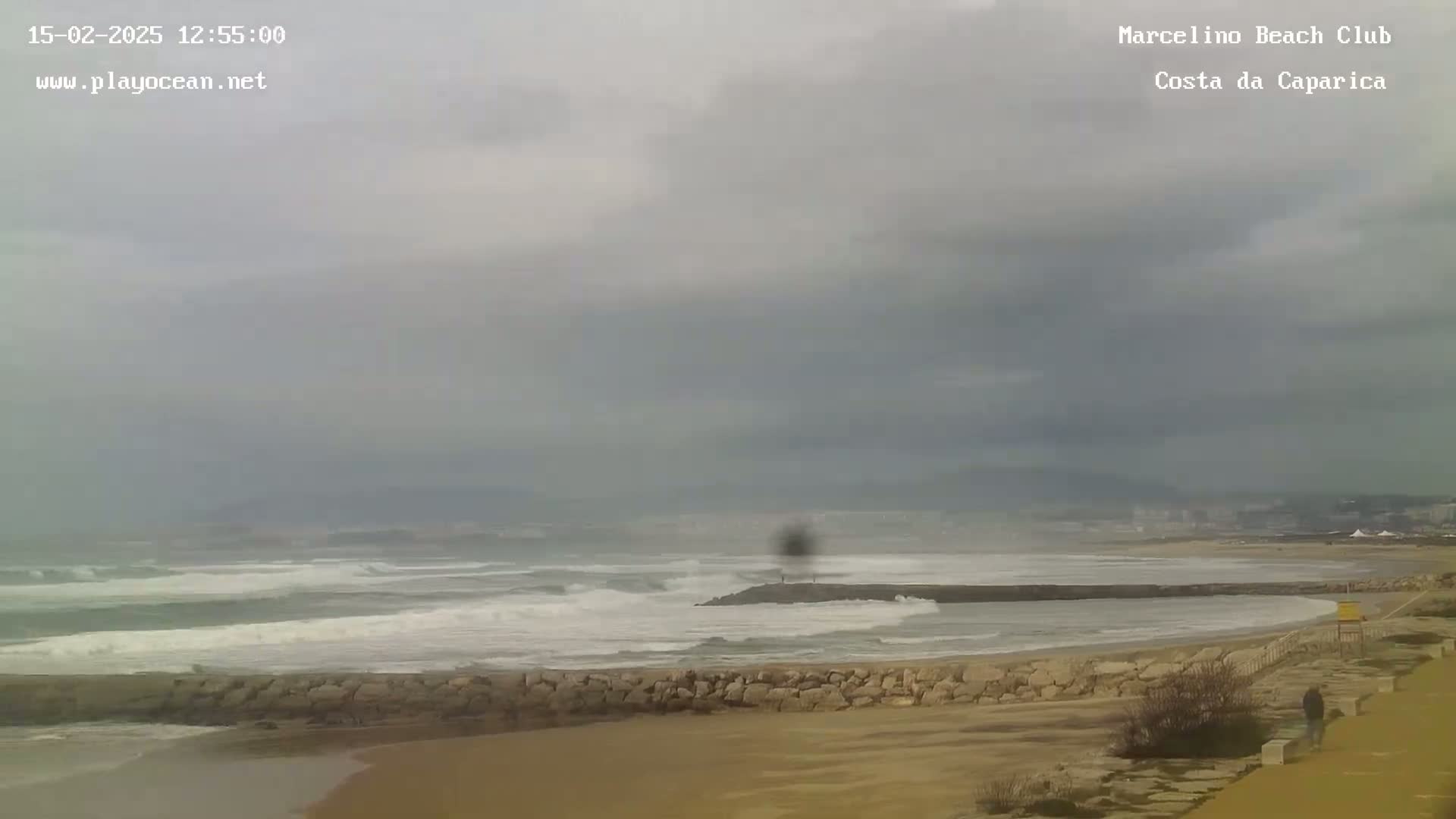 A sandy beach with a rocky breakwater stretches into a gray, overcast sky above a wavy ocean.