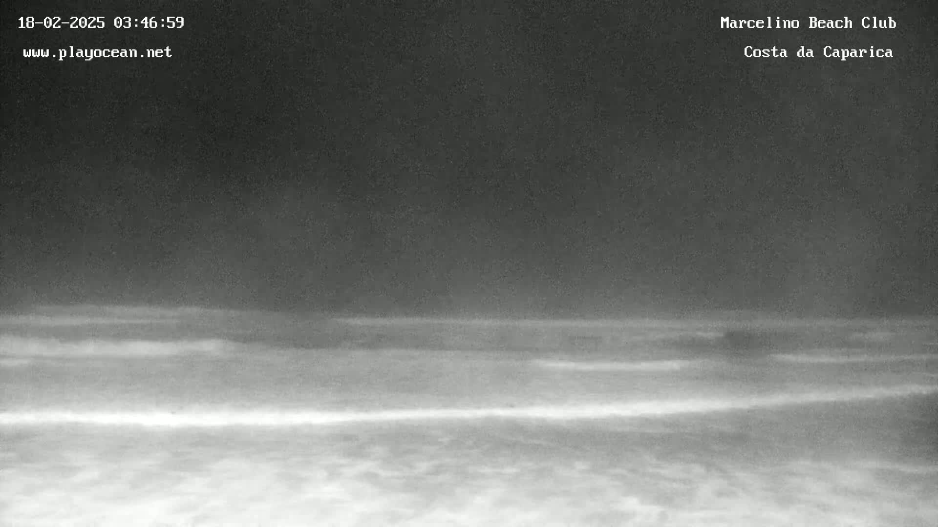 A nighttime beach scene shows dark, calm water meeting a dark sandy shore under a cloudy sky.