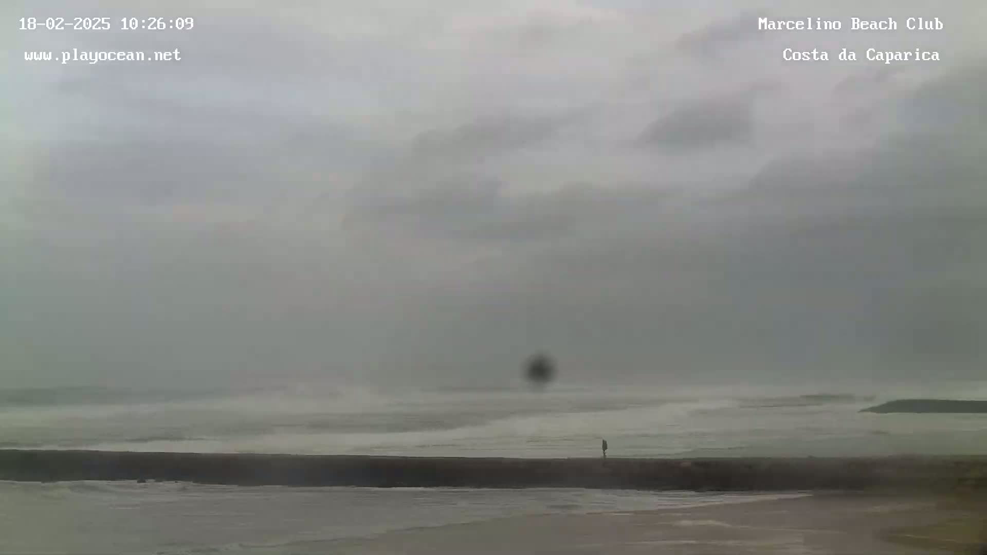 A person stands on a seawall overlooking a choppy ocean under an overcast sky.