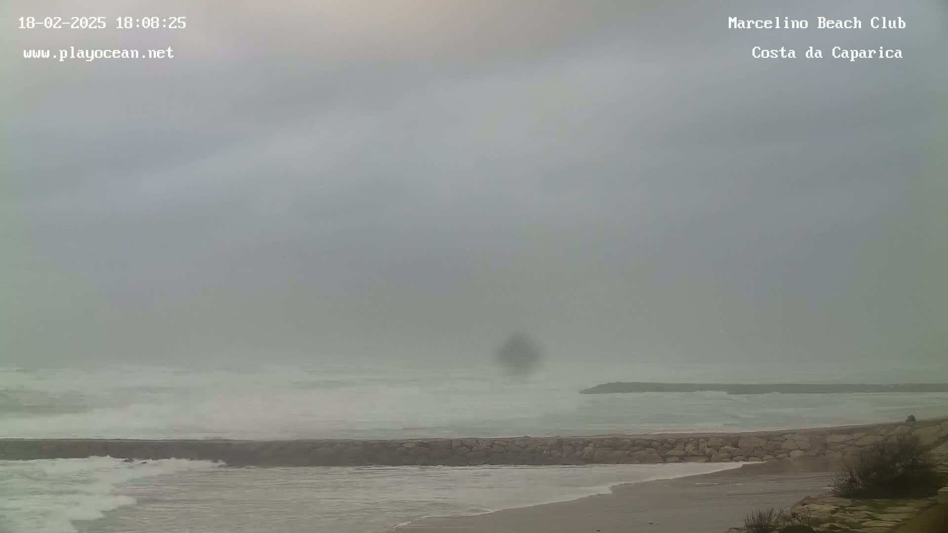 A low-visibility, overcast day shows a rocky breakwater separating a beach from choppy ocean waves.