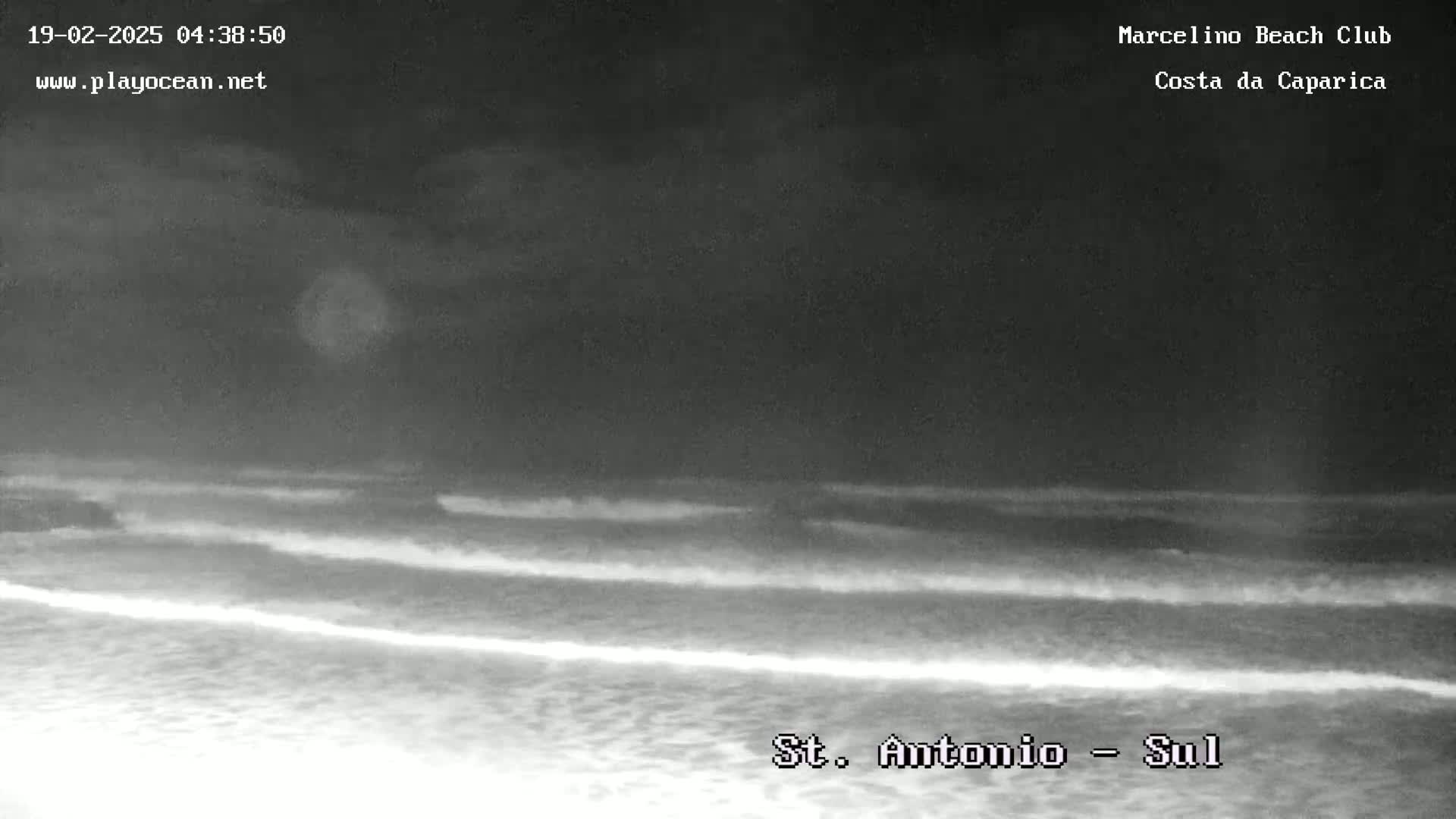 A nighttime, low-light image shows a dark beach with gently rolling waves under a cloudy sky.