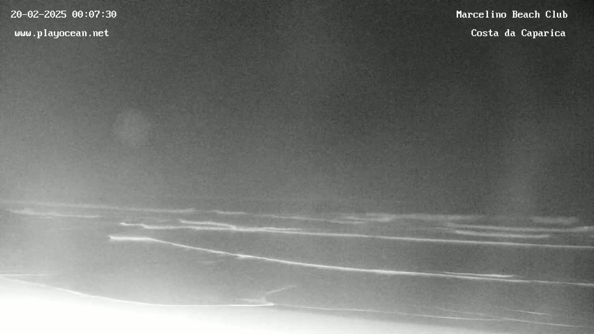 A nighttime, low-light image shows dark ocean waves lapping a sandy shore under a cloudy sky.