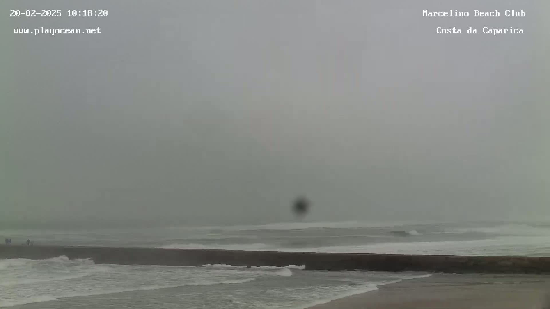 A hazy, overcast day shows a beach with a long, dark jetty, waves breaking against it, and a few people walking along the shore.