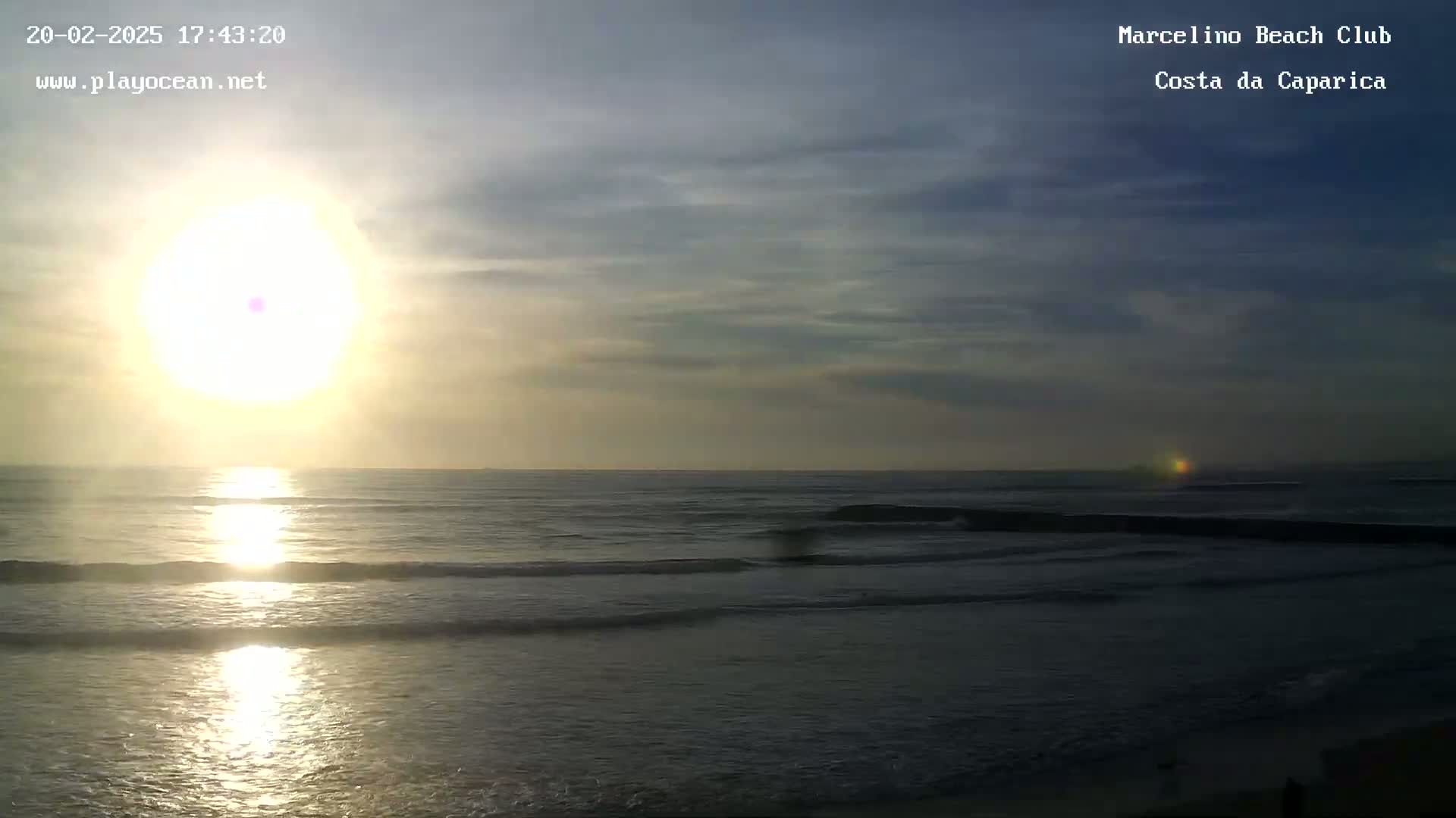 The setting sun shines over a calm ocean with small waves under a partly cloudy sky.