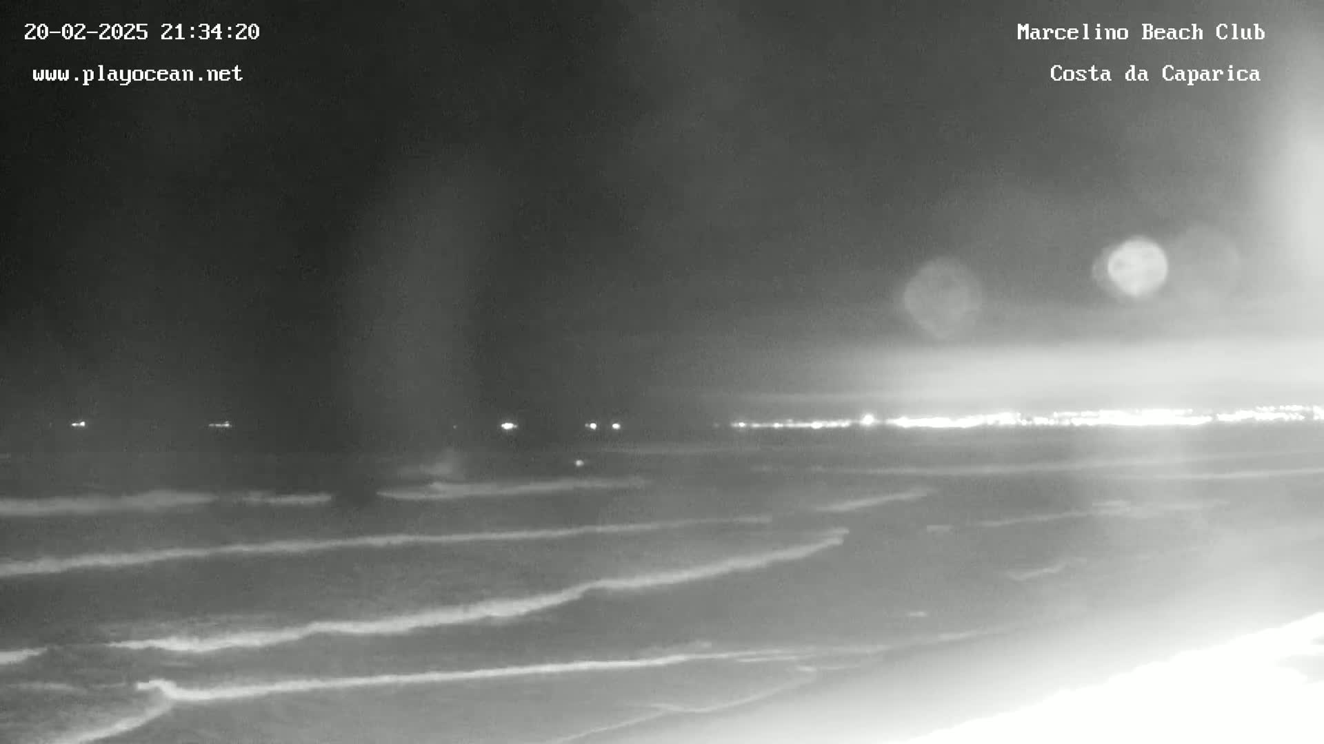 A nighttime view of a beach with gently rolling waves under a dark sky, showing a distant line of lights on the horizon.