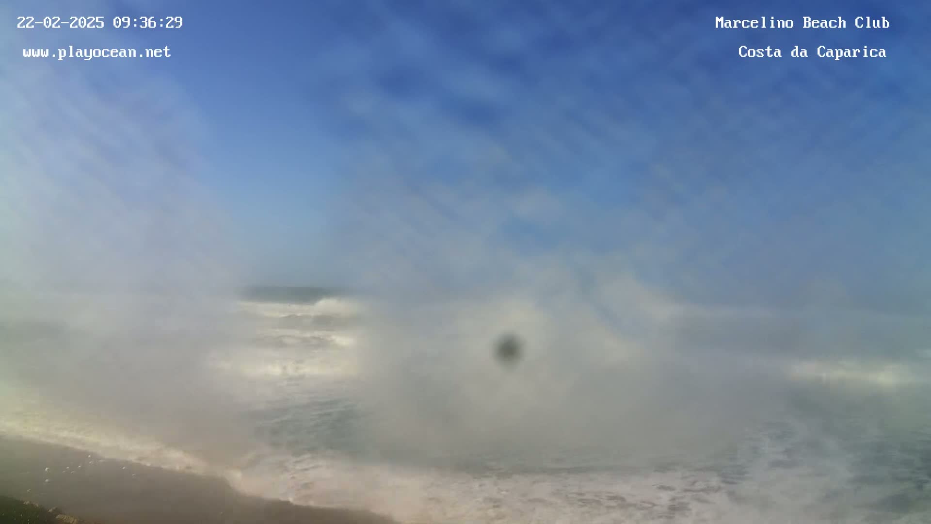 A hazy, misty ocean scene shows waves crashing on a sandy beach under a mostly clear blue sky.