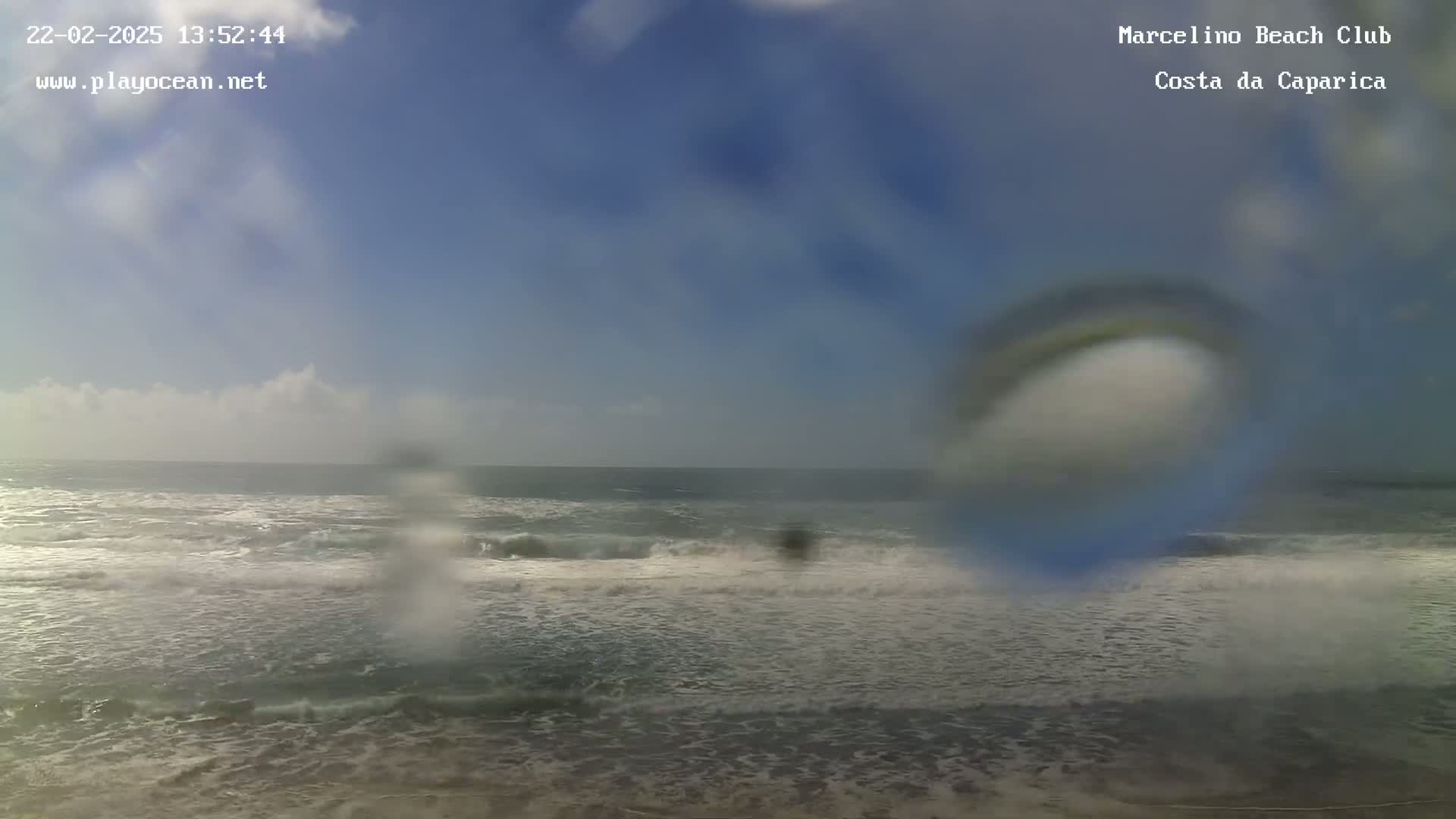 A mostly sunny day shows a beach with moderate waves, viewed through a rain-covered lens.