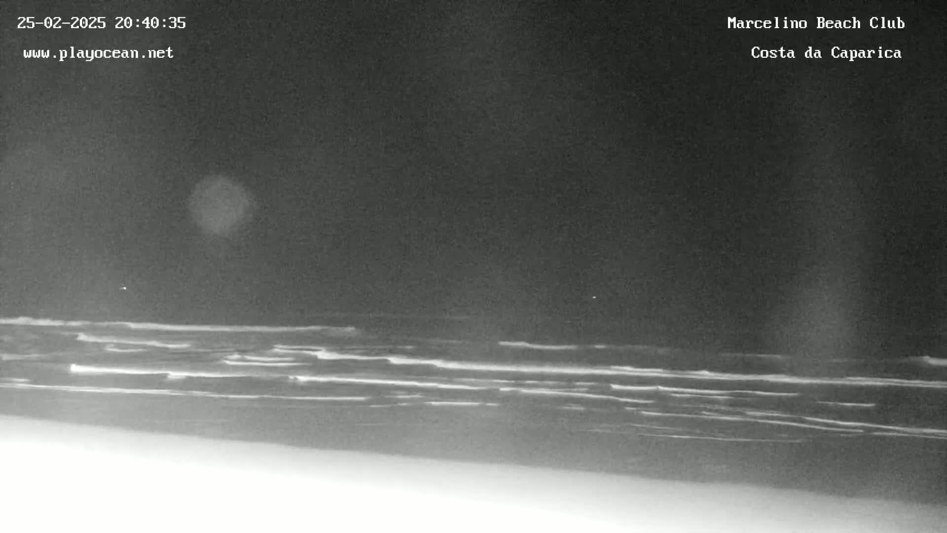 A nighttime, low-light view shows dark waves rolling onto a beach under a dark sky.