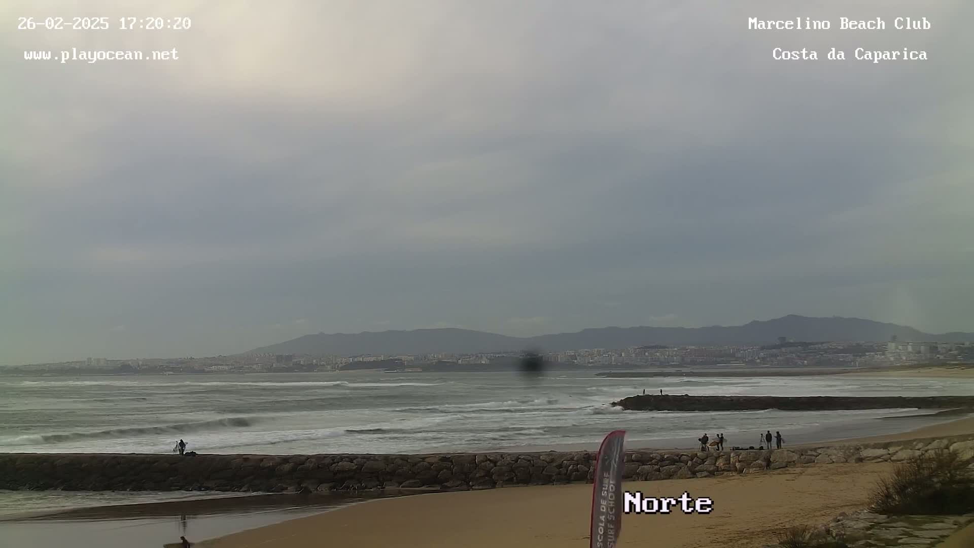 A beach with a rocky jetty, some people, and choppy waves under an overcast sky, with mountains and a distant city in the background.
