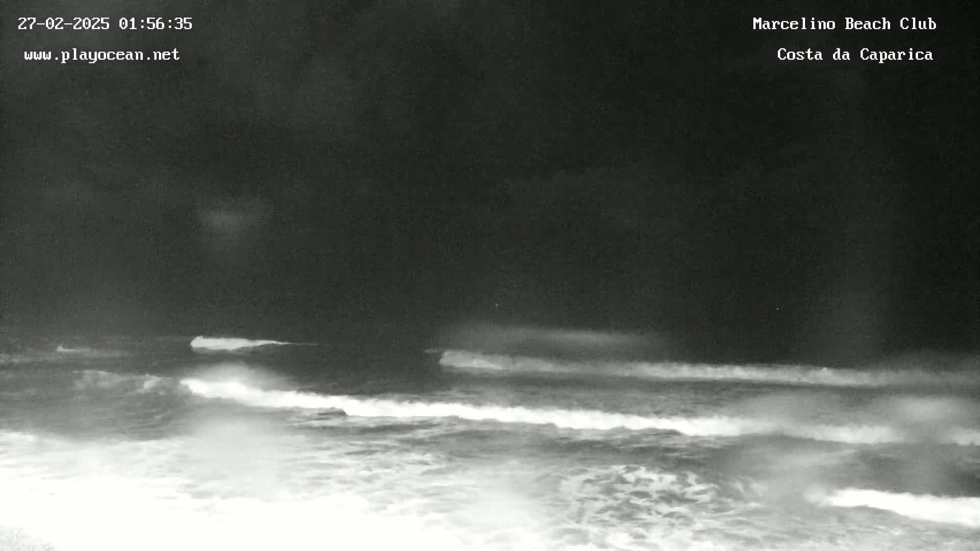 A nighttime, low-light image shows ocean waves rolling onto a shore under a dark sky.