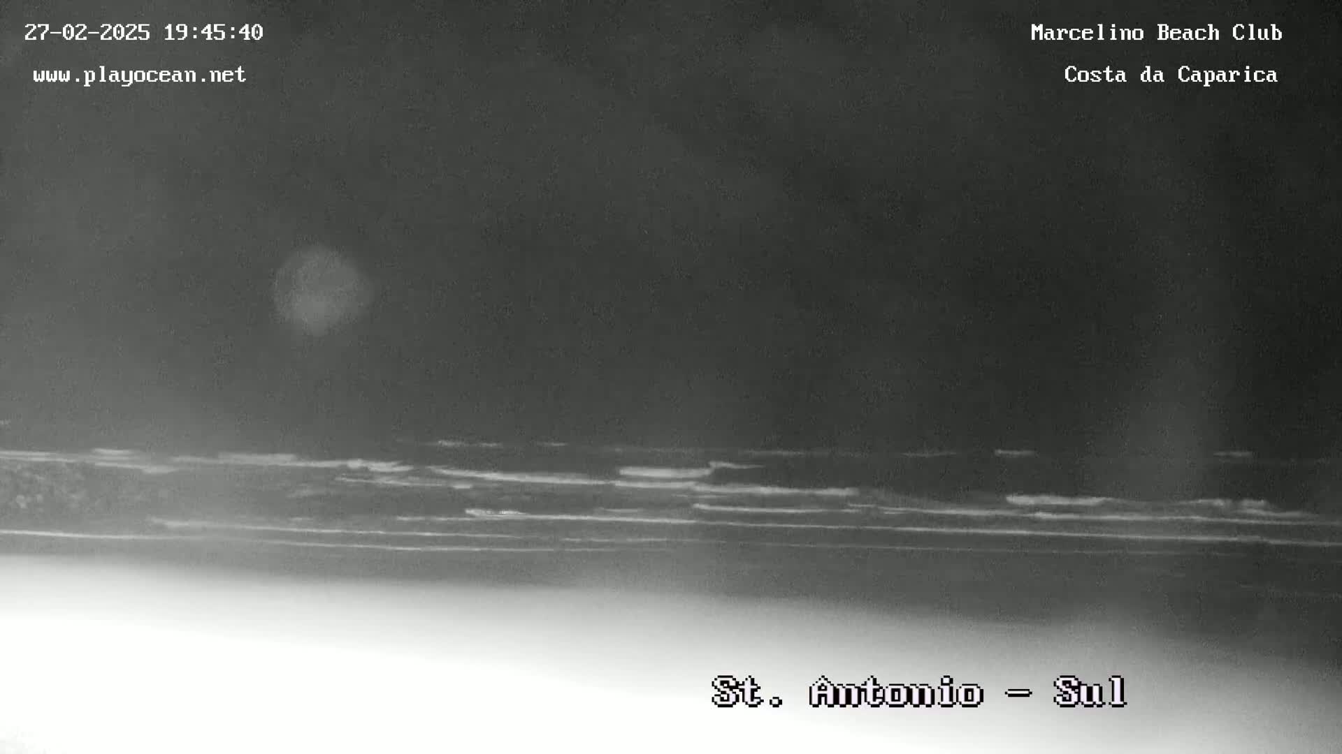 A nighttime, low-light view of a dark ocean with small waves breaking on a shoreline.