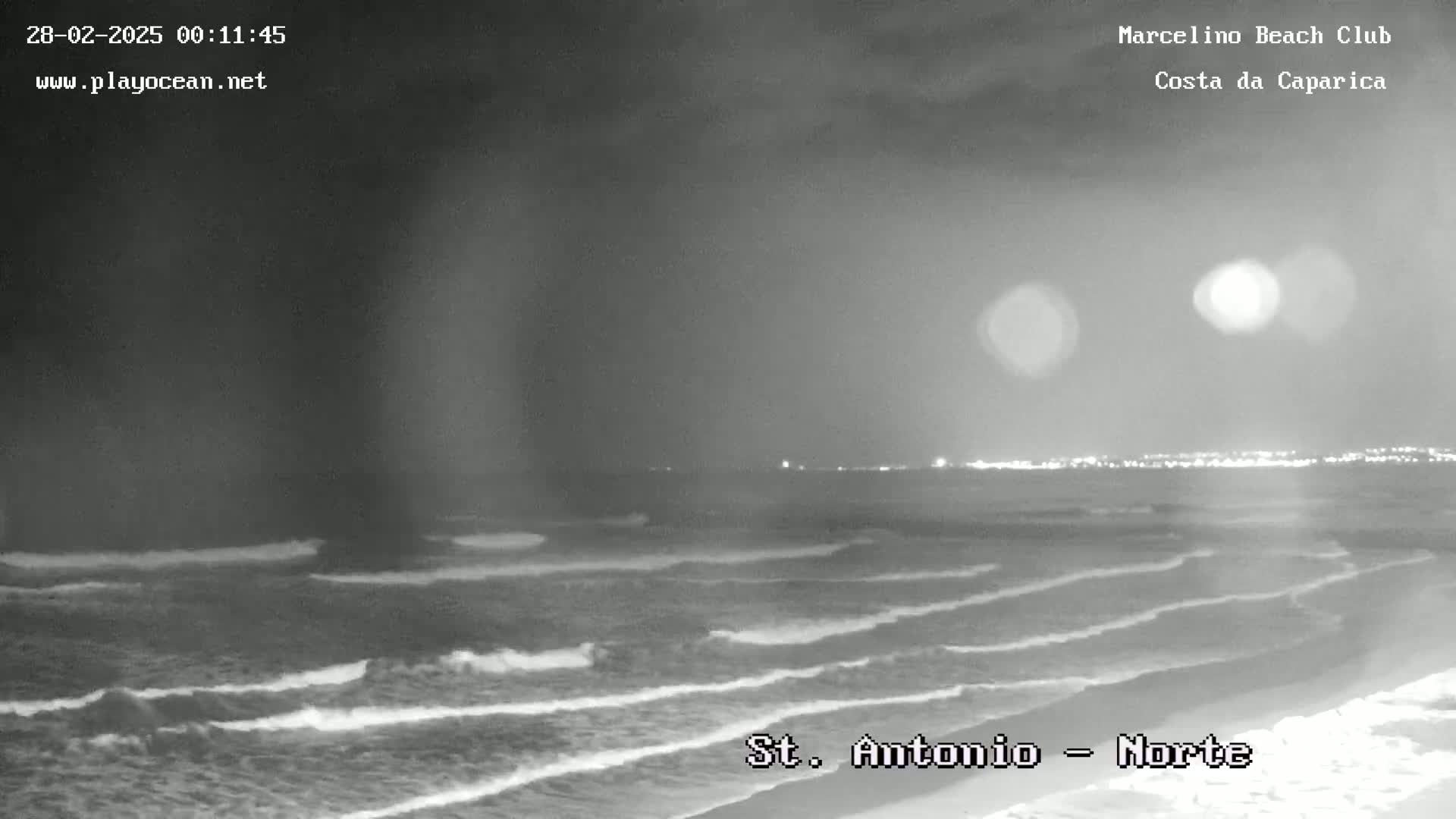 A nighttime, grayscale view of a beach with moderate waves rolling onto the shore under a dark sky, with distant city lights visible on the horizon.