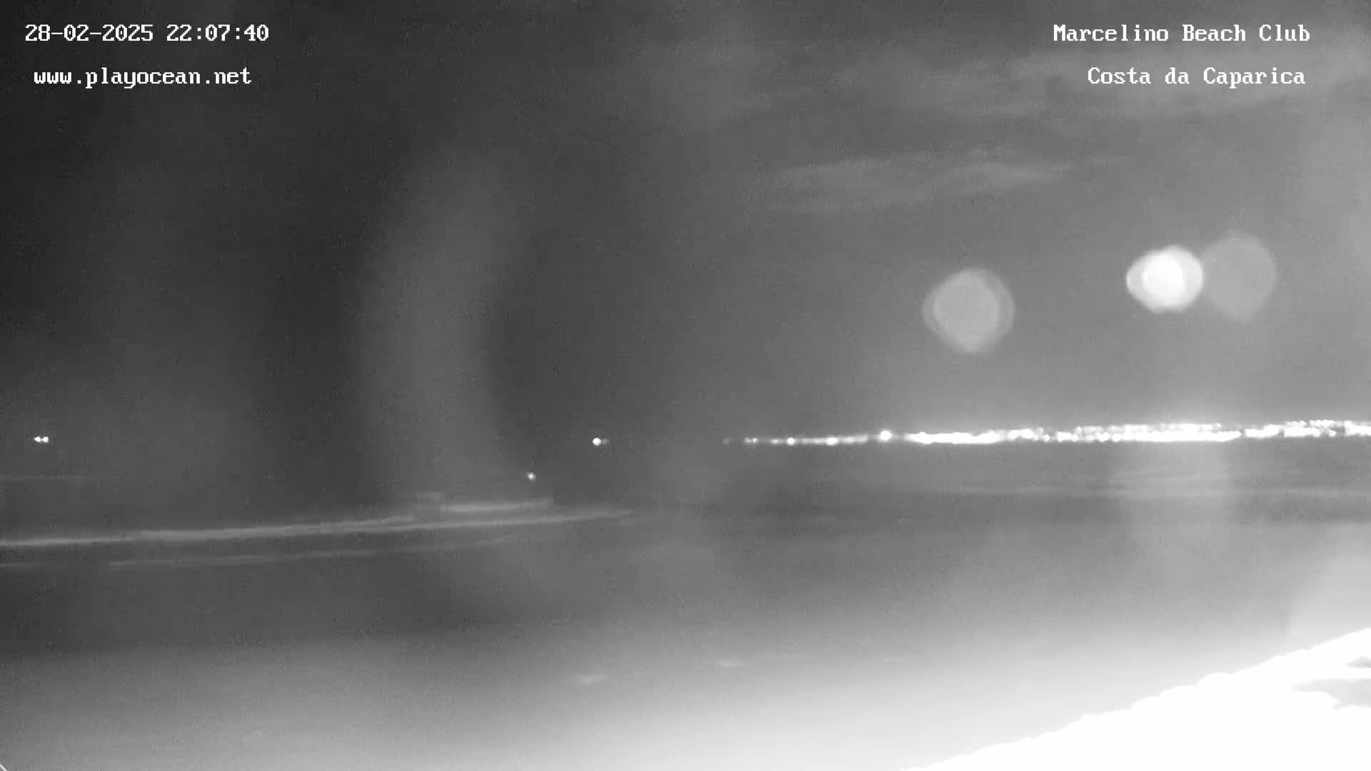 A nighttime, low-light view of a dark beach with a faintly visible city skyline in the distance.