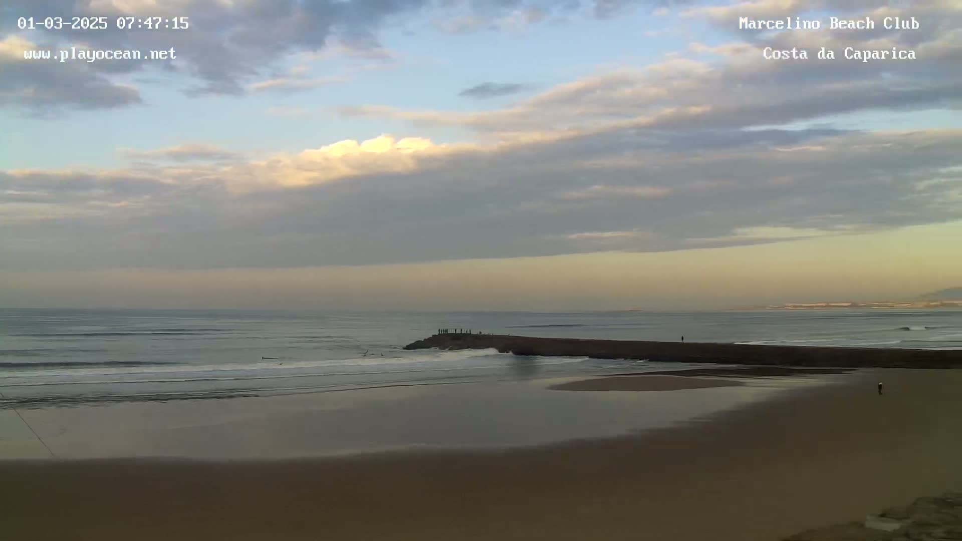 A mostly cloudy, pale sky overlooks a calm ocean beach with small waves breaking near a rock jetty where several people stand.