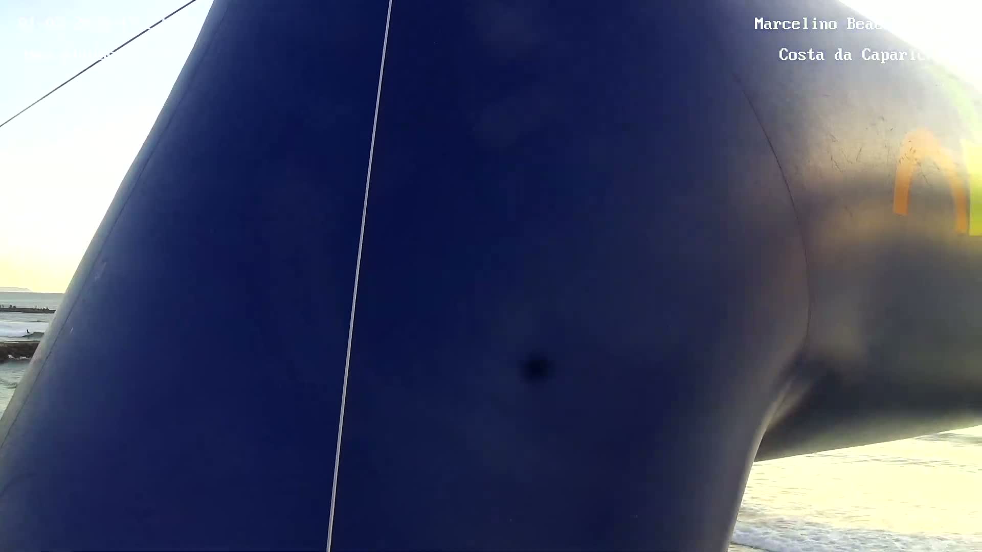 A close-up view shows a portion of a large, dark-blue inflatable structure near an ocean under sunny skies.