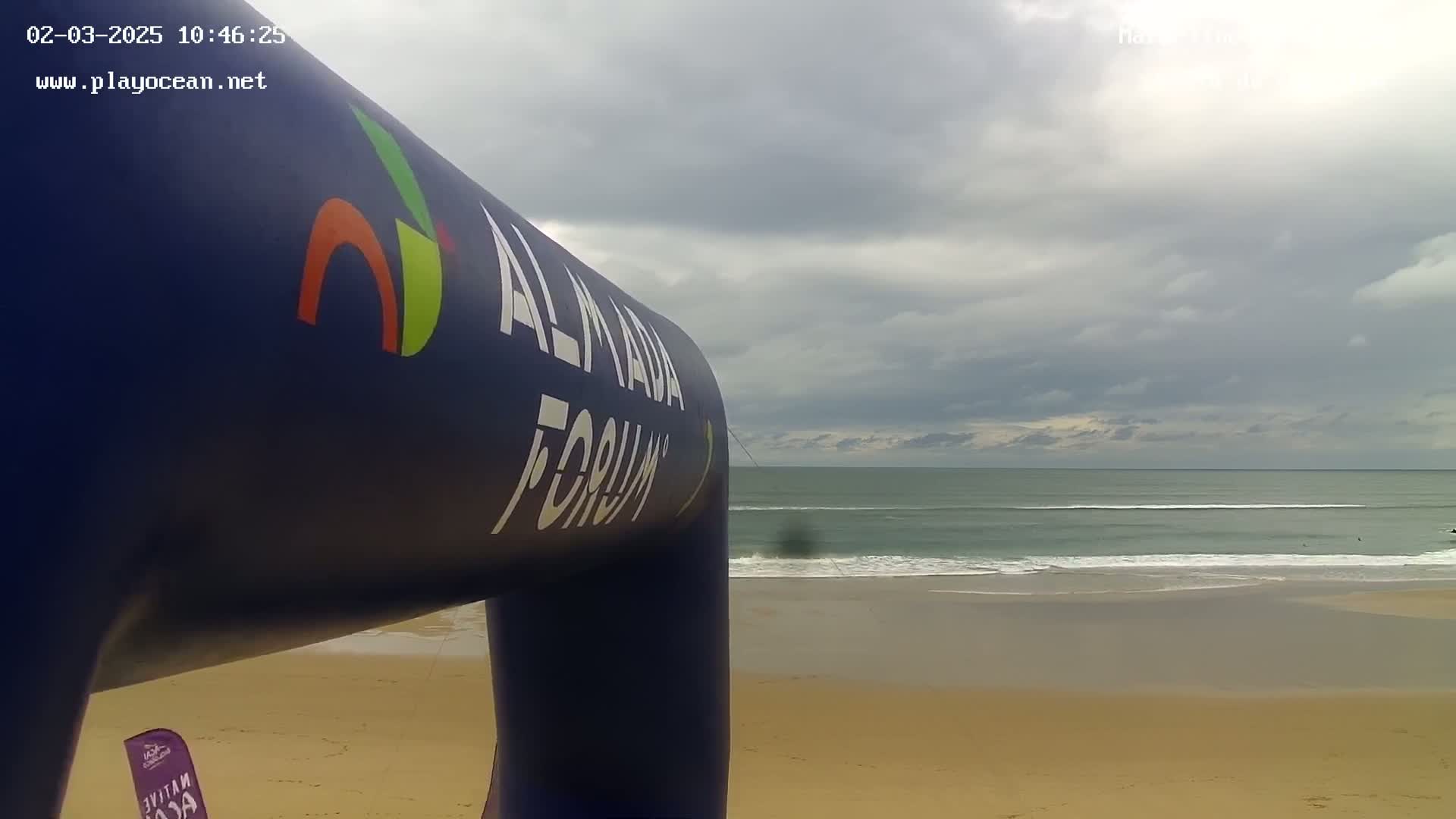 A large inflatable archway stands on a sandy beach overlooking a calm ocean under a cloudy sky.