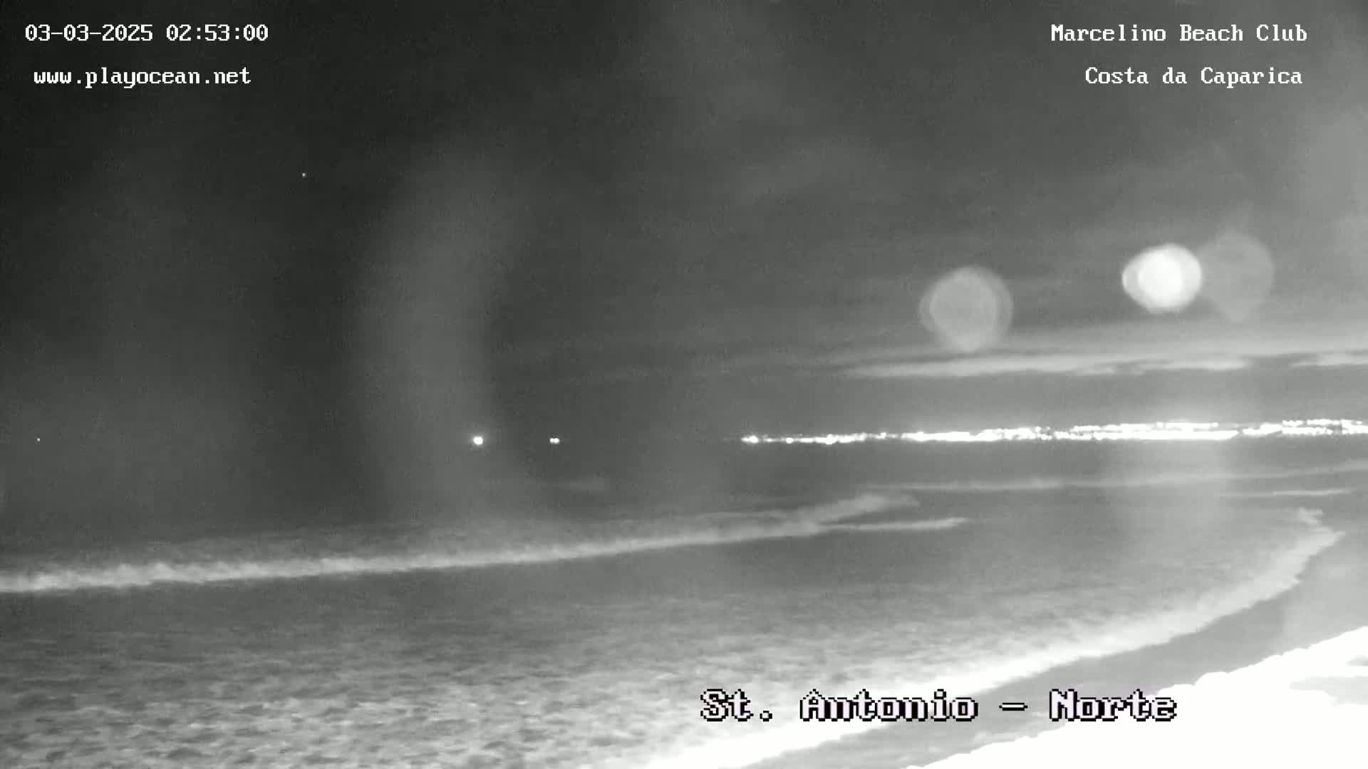 A nighttime, grayscale view of a beach with gently rolling waves under a mostly clear sky.