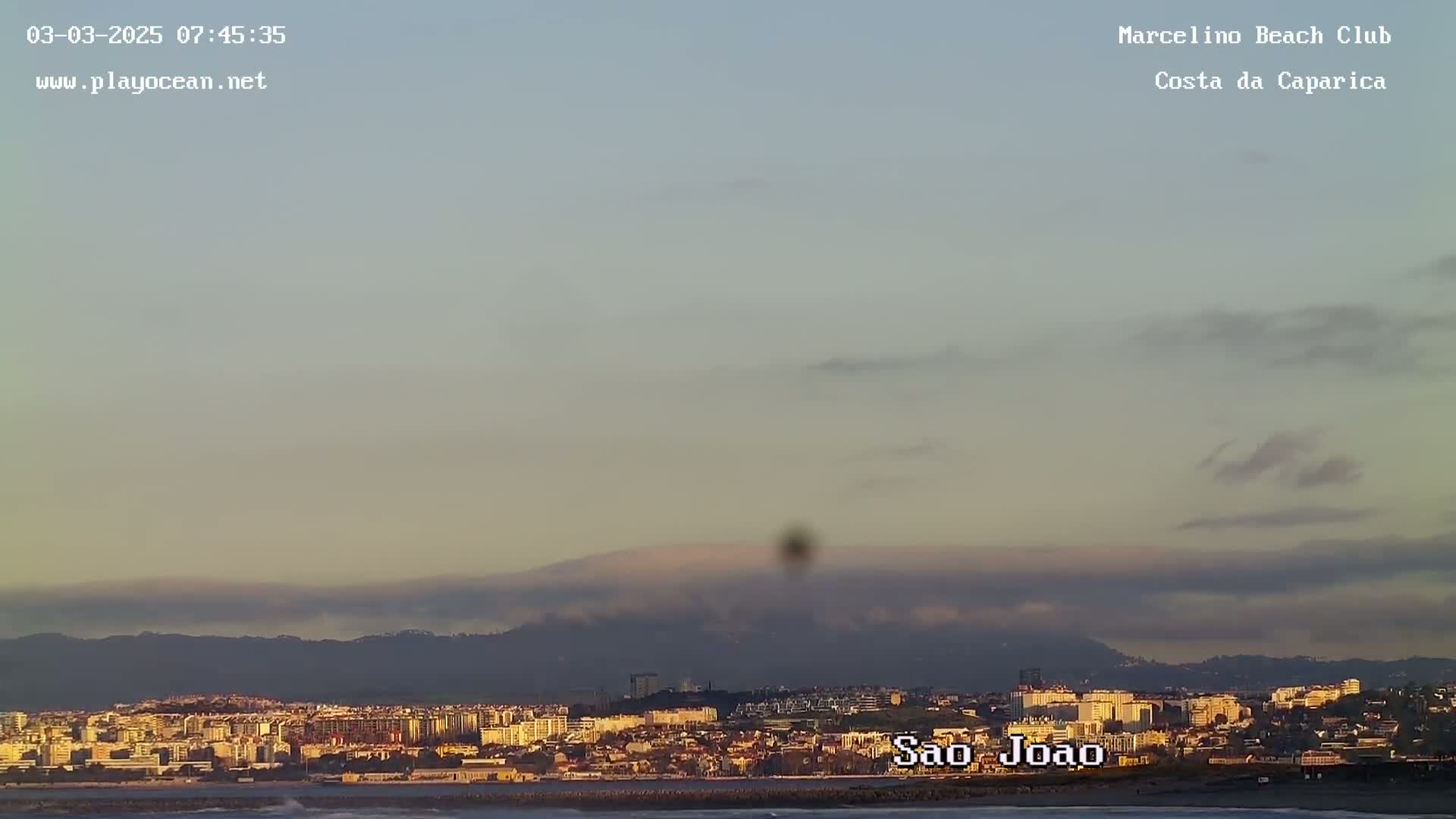 A coastal city sits under a hazy, pale sky, with mountains visible in the distance.