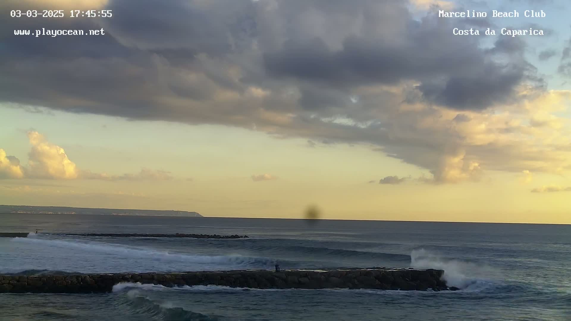 A partly cloudy sunset over a rocky ocean coast with waves breaking against a low wall, and a few people visible on the wall.
