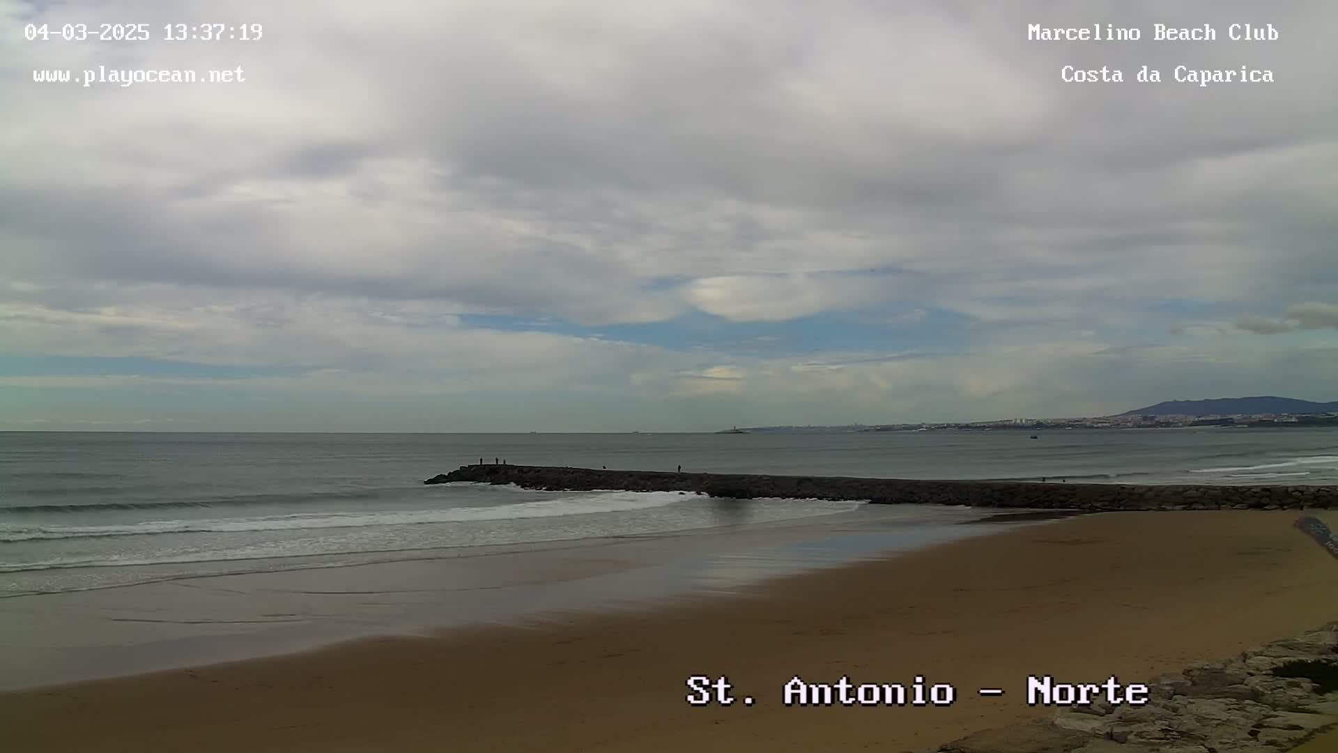 A sandy beach with a long, low rock jetty stretches into a calm ocean under a mostly cloudy sky.
