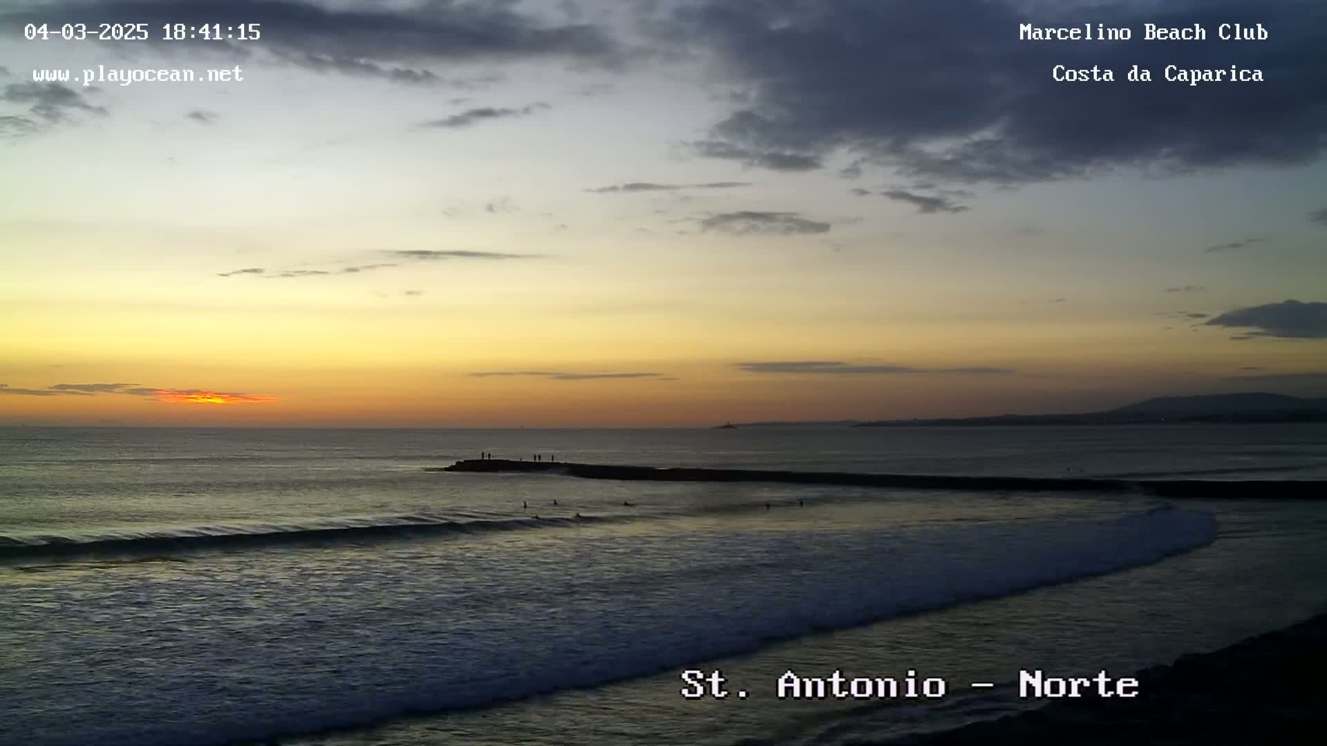 A sunset over a dark ocean with small waves, several people standing on a rock jetty, and a few surfers in the water.