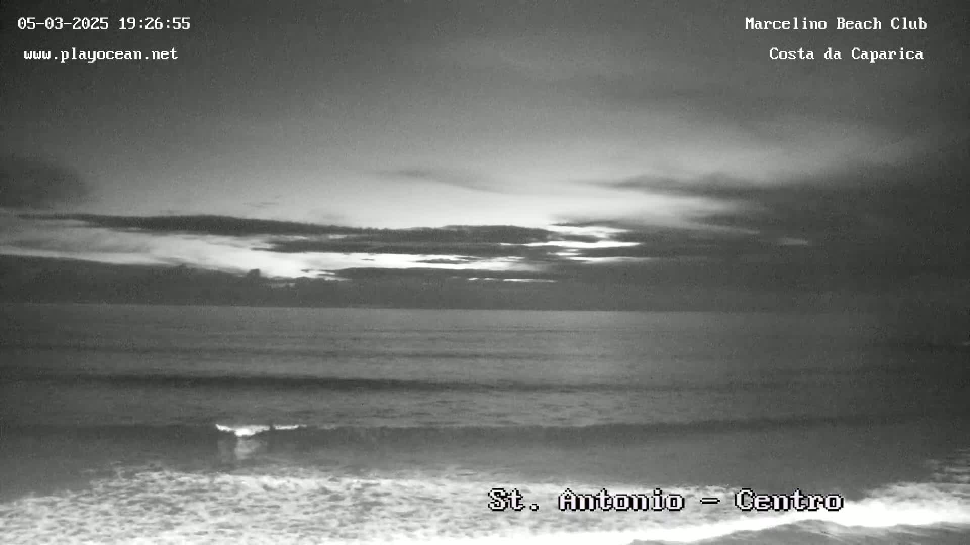 A grayscale image shows an ocean beach under a cloudy, twilight sky.