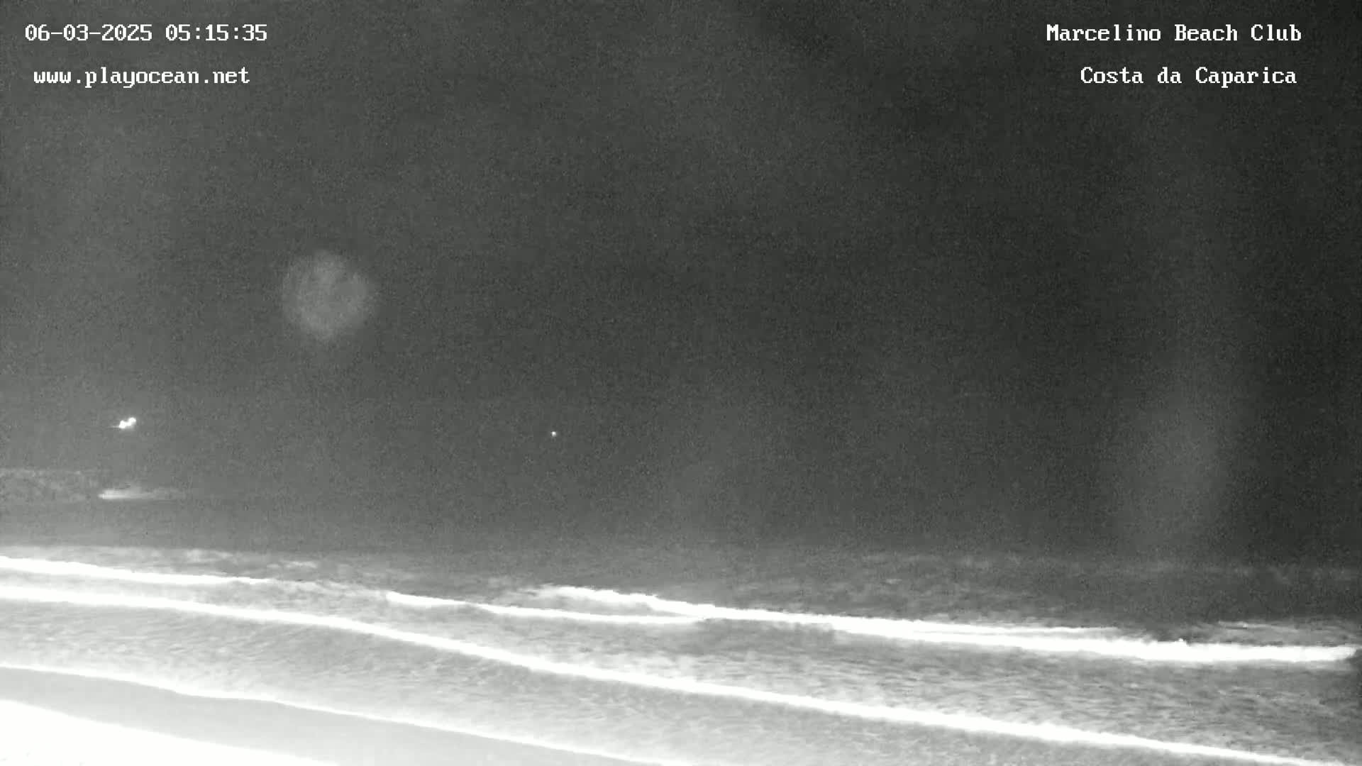 A nighttime, long shot of gentle waves lapping on a dark beach under a cloudy sky.