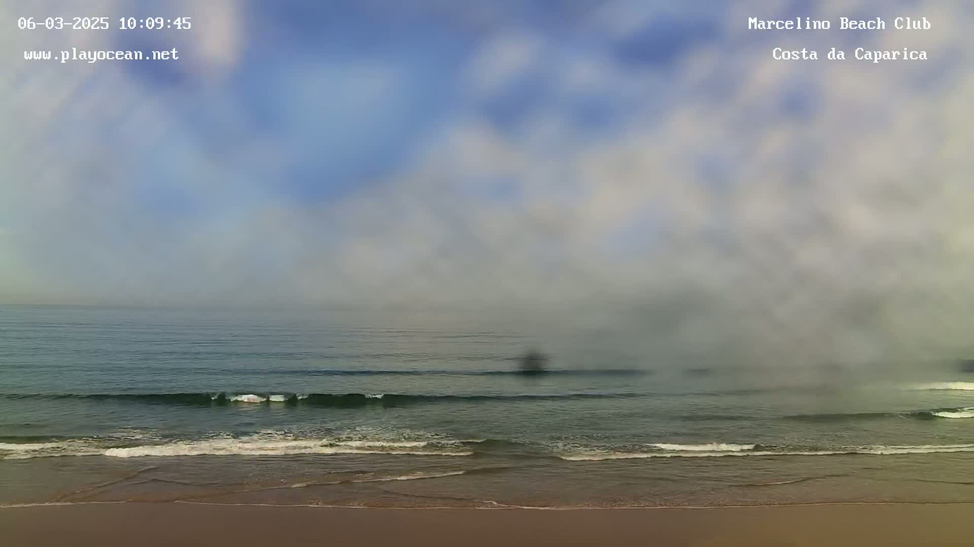 A hazy, overcast sky hangs over a calm ocean with gentle waves lapping a sandy beach.