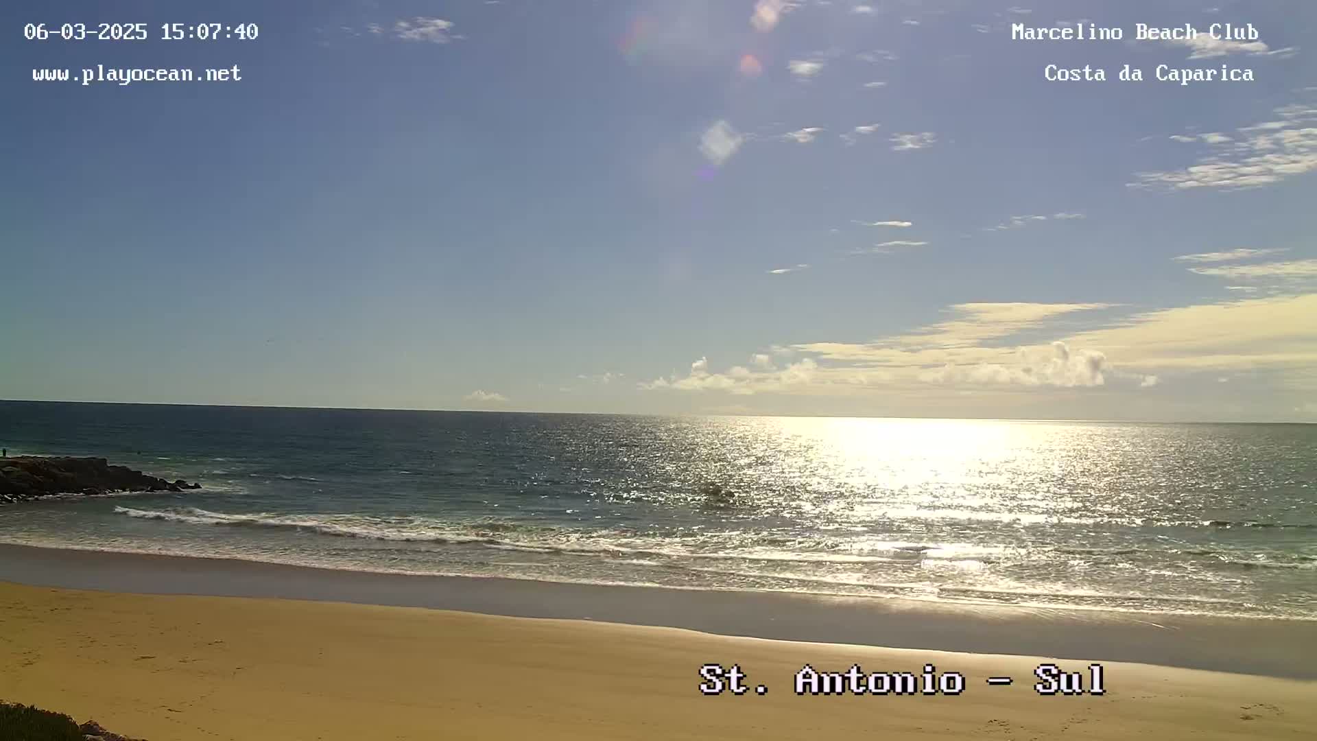 A sandy beach meets a calm ocean under a mostly sunny sky with a few scattered clouds.