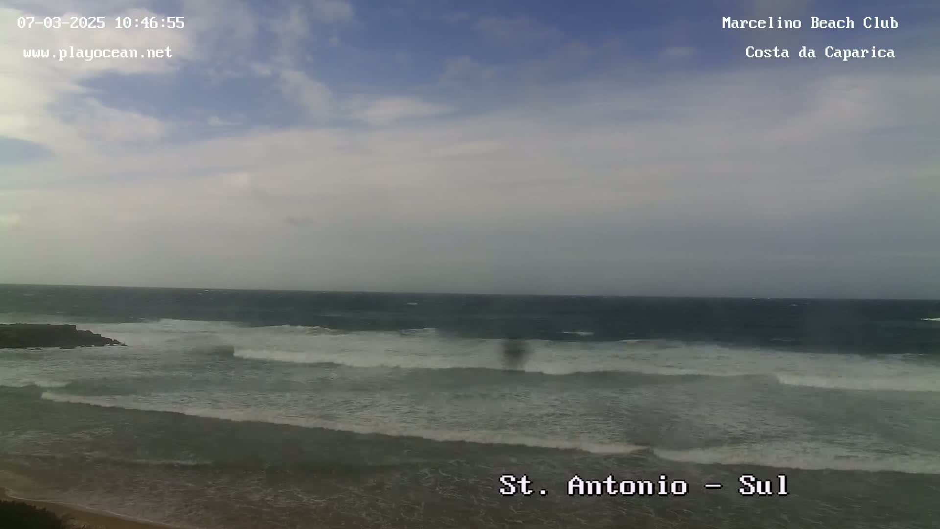 A partly cloudy day shows a wide expanse of ocean with moderate waves breaking on a sandy beach.