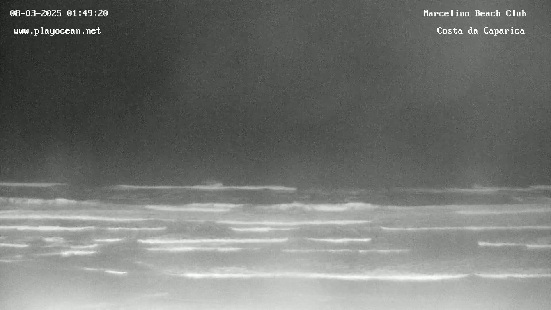 A nighttime, low-light image shows a dark ocean with small waves breaking on the shore under a dark sky.