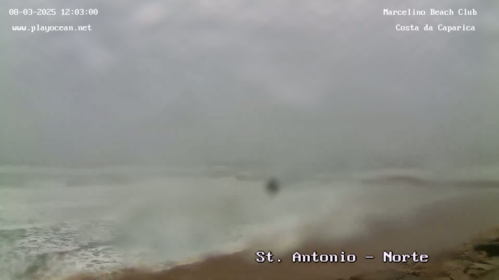 A hazy, overcast day shows a beach with turbulent waves crashing on the shore.