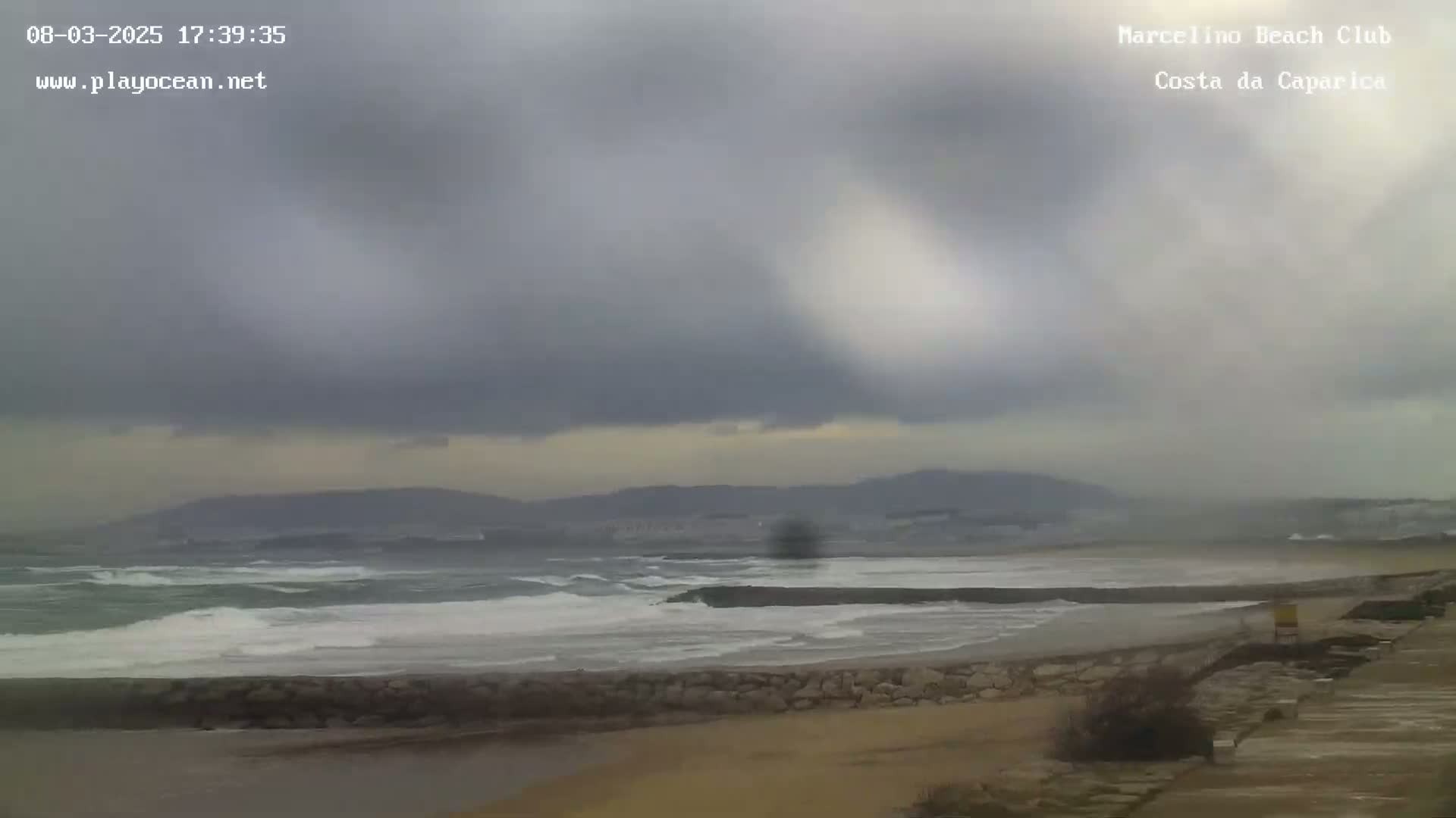 A partly cloudy day shows a beach with choppy waves and a distant mountain range under an overcast sky.