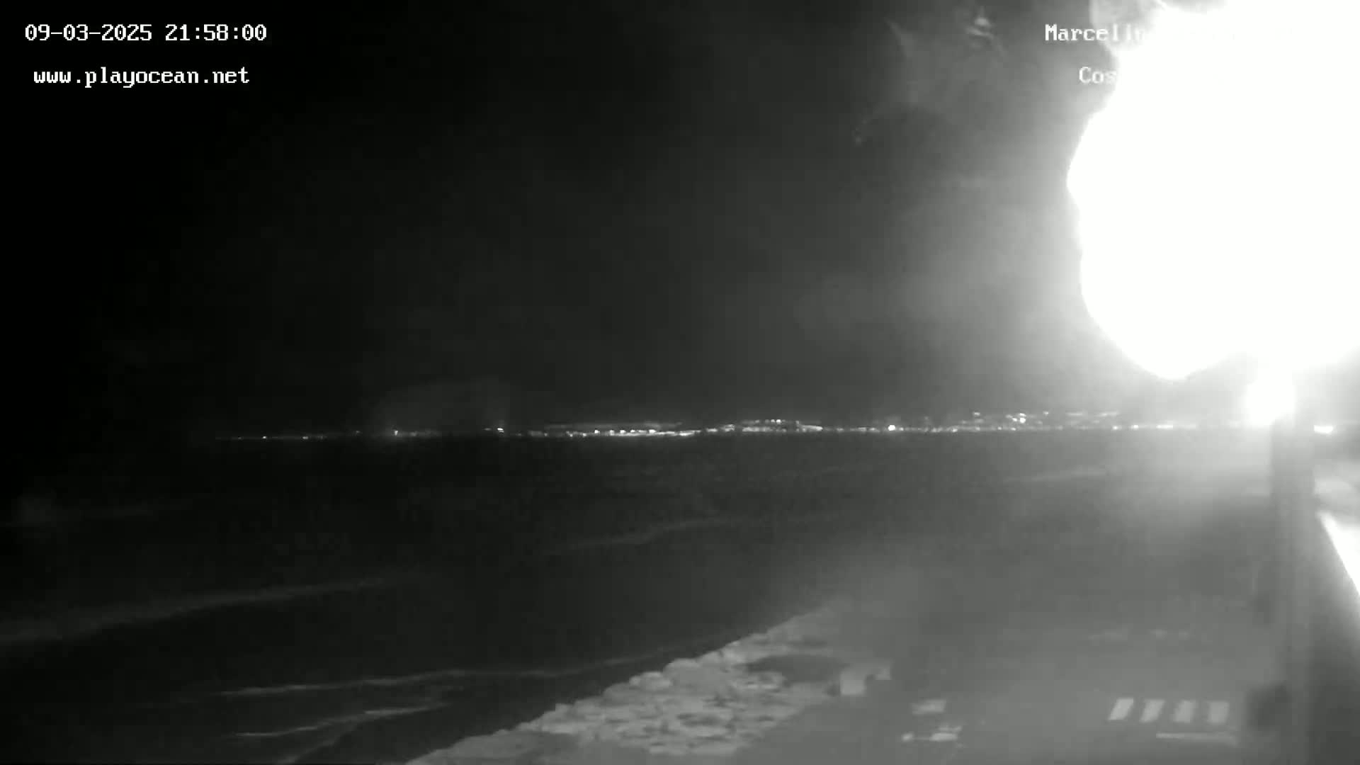 A nighttime coastal scene shows a bright light source in the upper right corner, with a dark body of water in the foreground and a faintly lit cityscape in the distance.