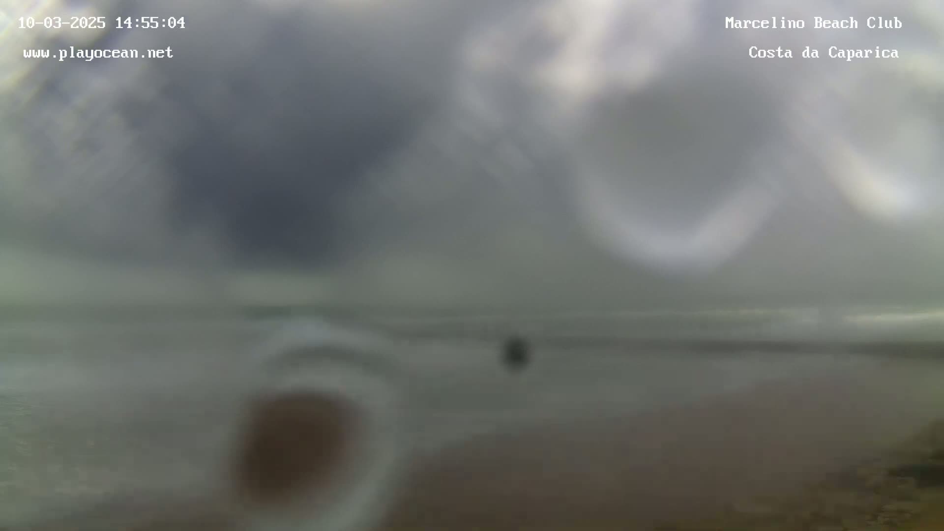 A blurry image shows an overcast sky over a calm ocean with a dark beach in the foreground.