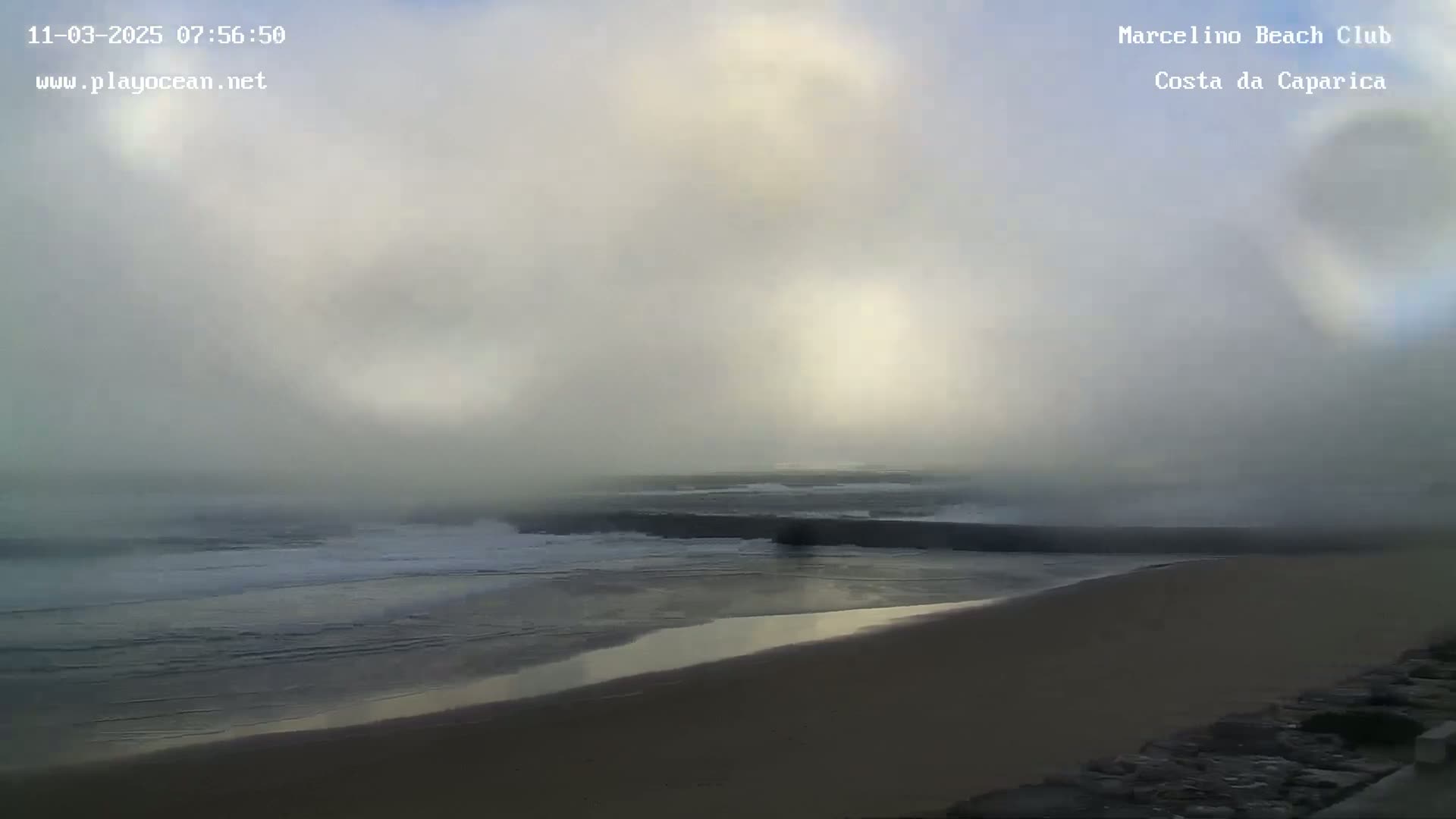 A foggy morning reveals a beach with waves breaking near a dark structure extending into the ocean.