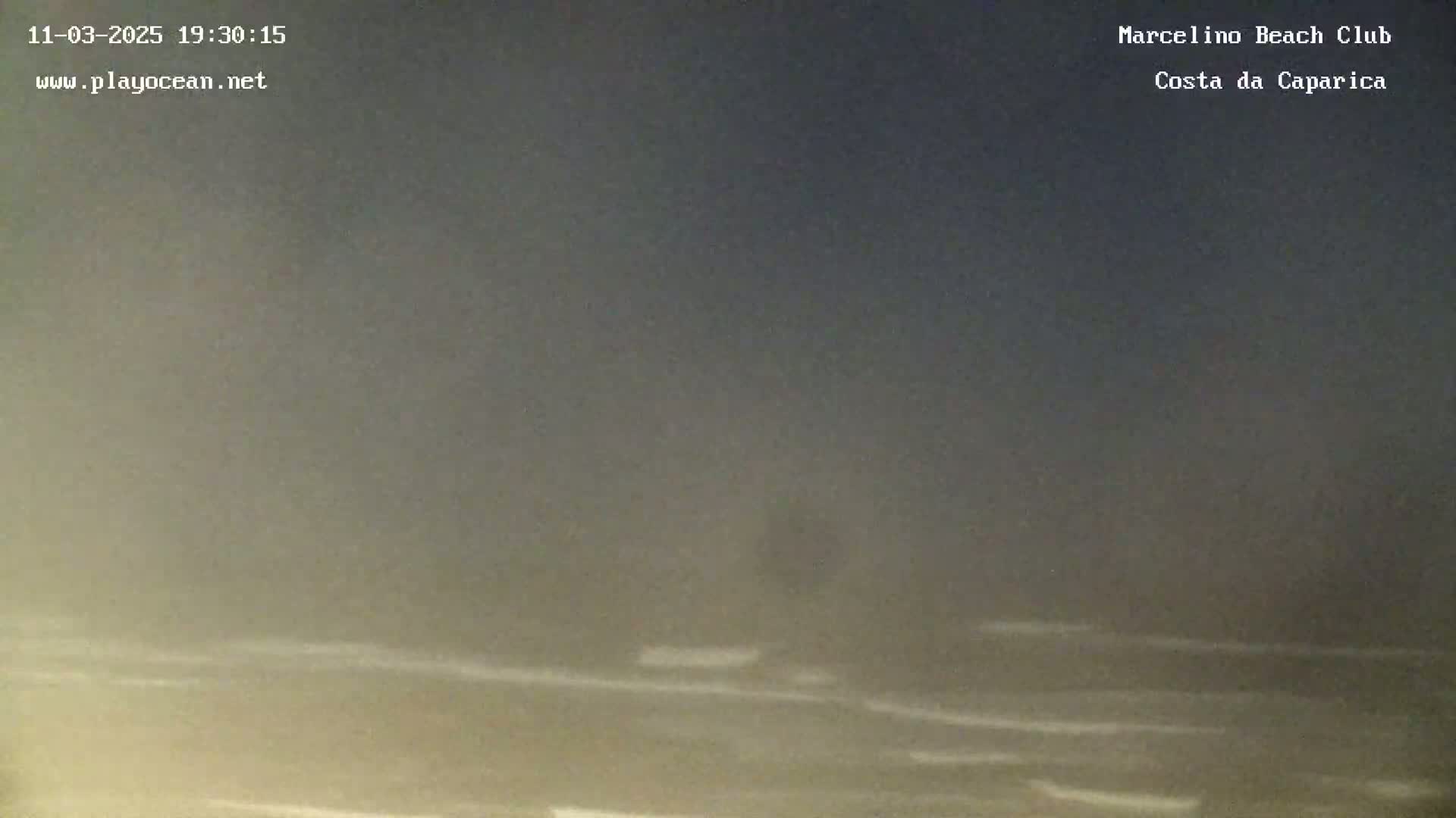 A dark, hazy beach scene at night shows a dimly lit ocean with small waves under a dark sky.