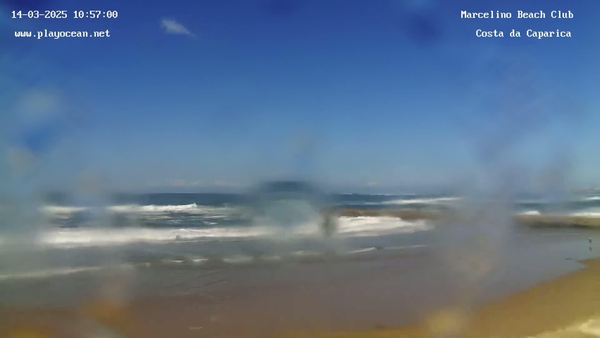 A hazy view shows waves breaking on a sandy beach under a clear blue sky.
