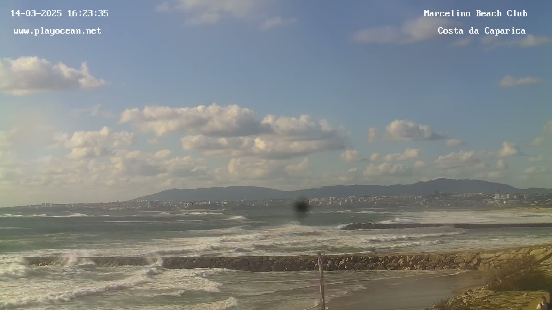 A partly cloudy sky overlooks a rocky coast with choppy waves crashing against a breakwater, and a distant cityscape is visible beyond the water.