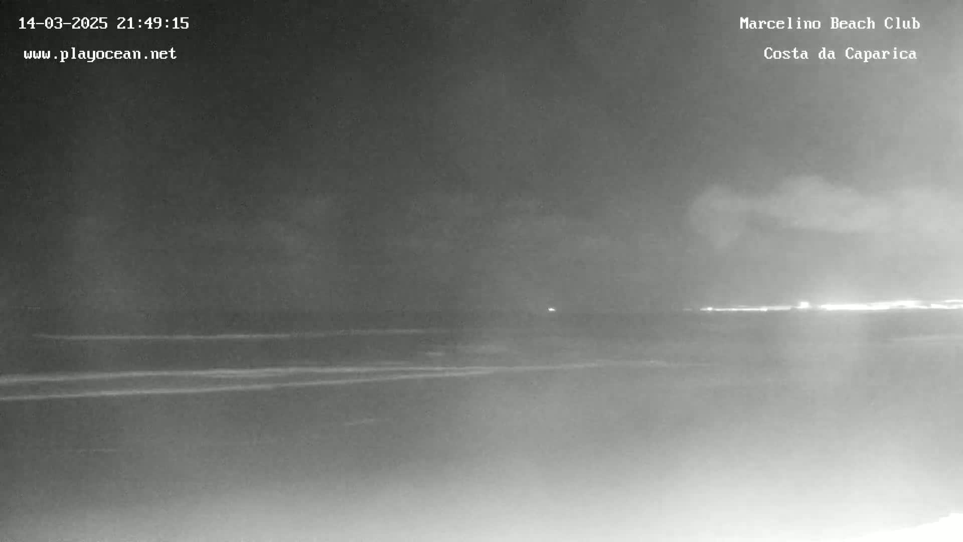 A nighttime, low-light view of a dark beach and ocean with a faintly visible shoreline in the distance.